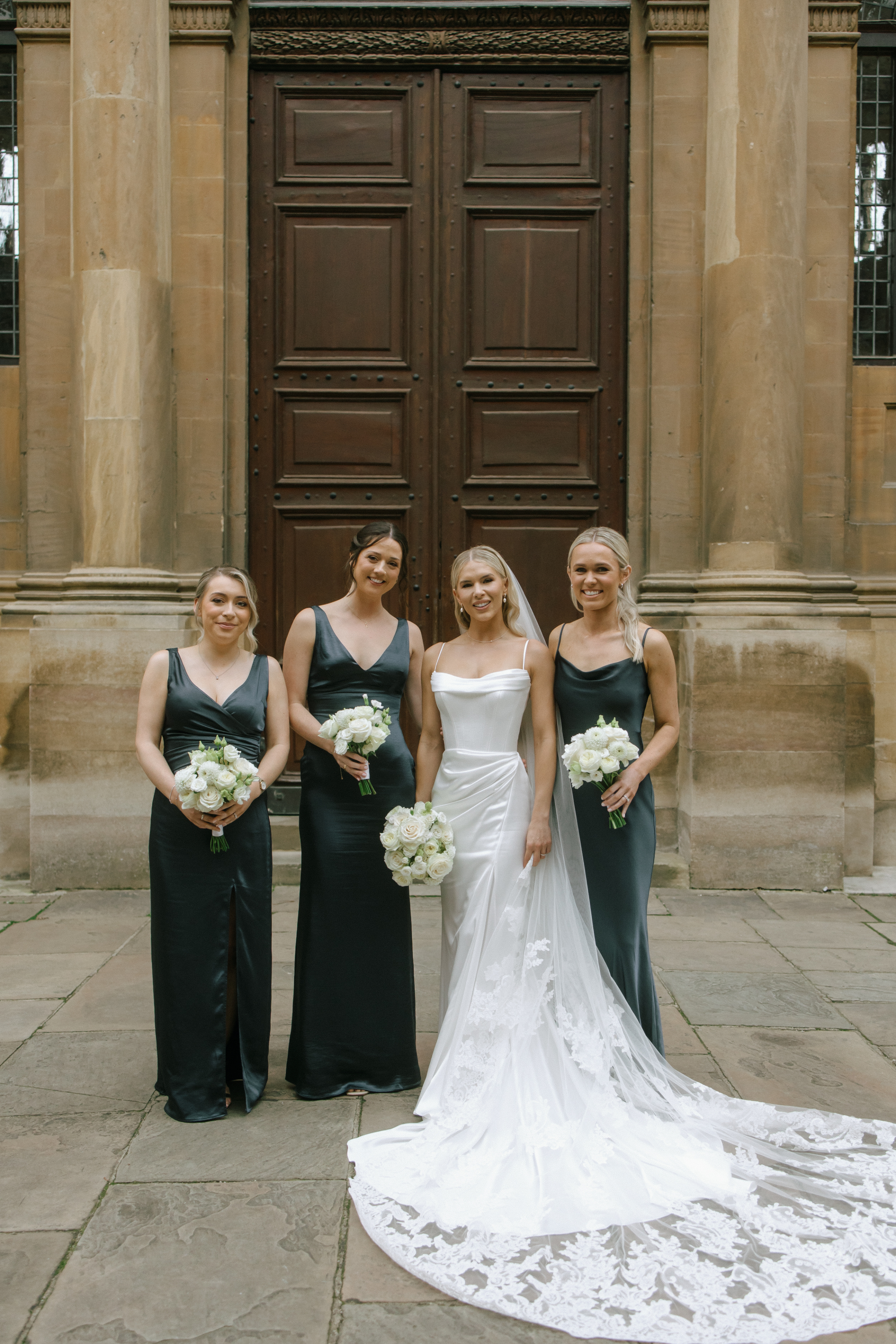 A Classy & Modern City Wedding at the Bodleian Library With Jamaican-Guyanese Heritage 44 The bride stands with her three bridesmaids in front of grand wooden doors at the Bodleian Library, wearing a fitted satin gown with an embroidered veil, while the bridesmaids wear sleek slate satin dresses and hold white rose bouquets.