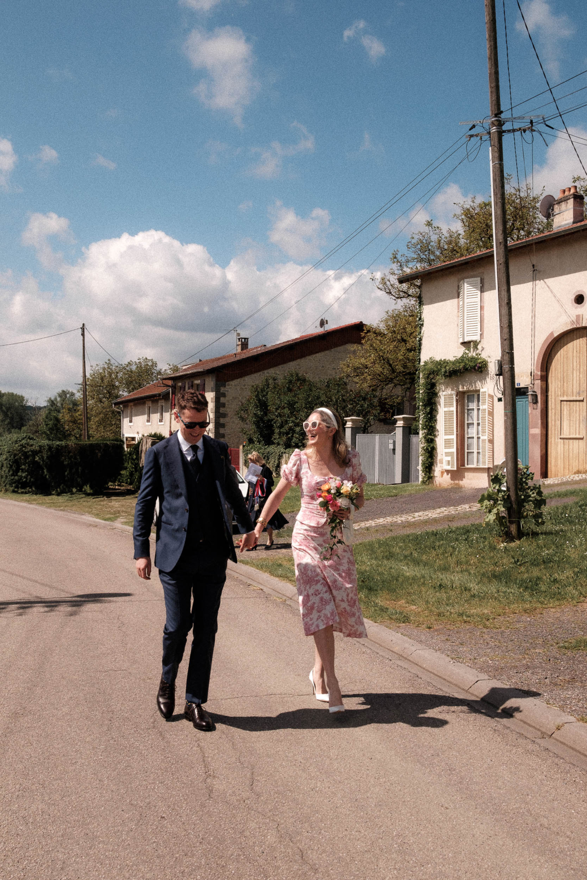 Constance and Nial walking hand in hand through the streets of Manonviller, moments after their ceremony. A joyful, sun-soaked image that captures the charm of an intimate French village wedding, with stylish non-traditional details.