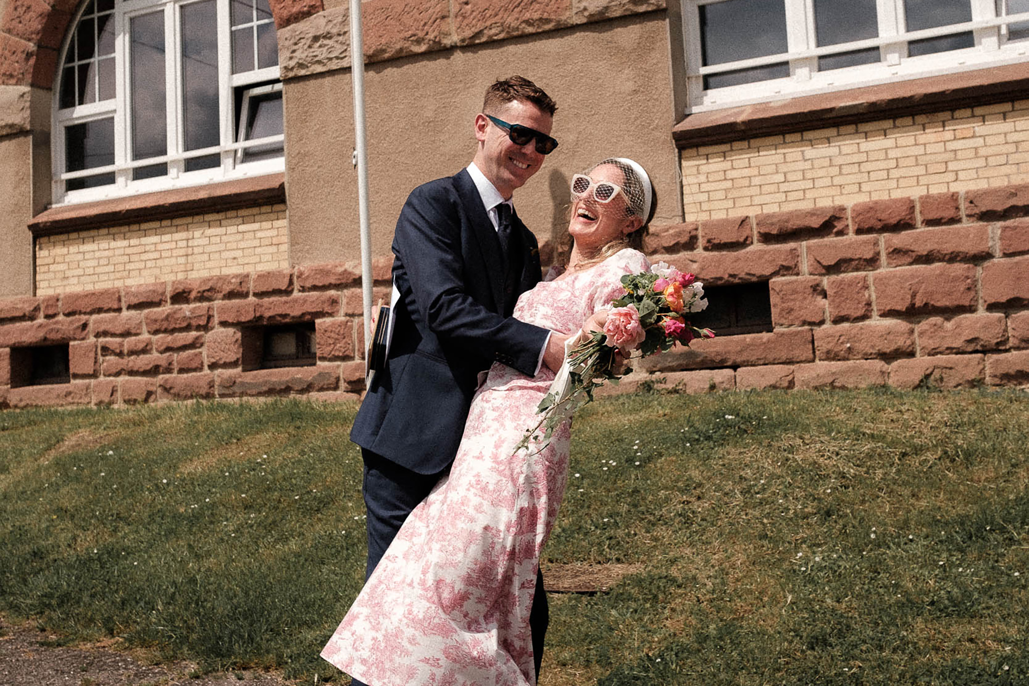 Groom holding his bride in a romantic gesture as she leans back and laughs. She wears a non traditional pink two-piece, sunglasses and a headband with birdcage veil. Intimate French village wedding