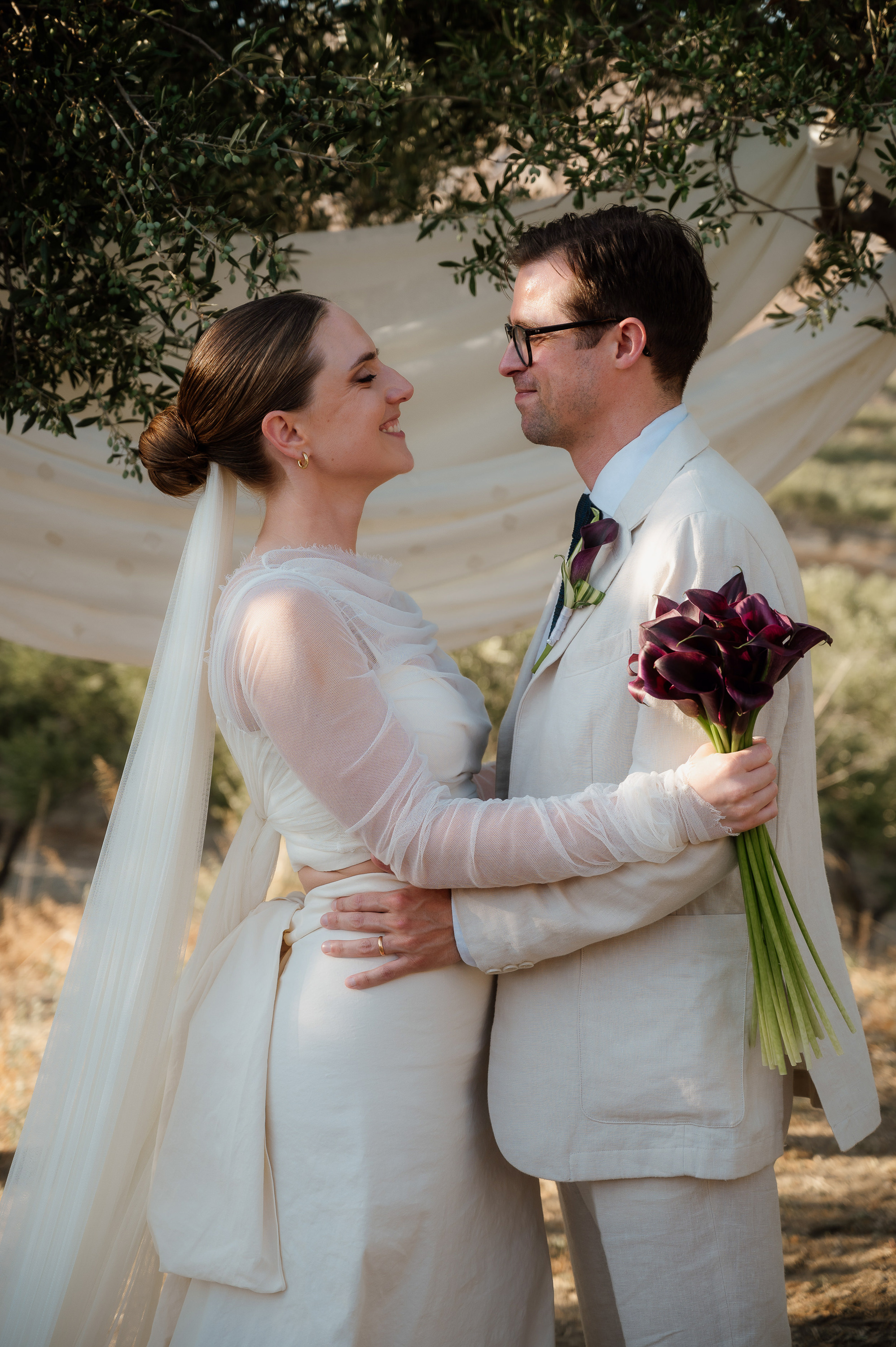 Alex & Jon’s Non-Traditional Wedding in the Olive Groves of Crete 5 Bride wearing a Cortana dress and AM Faulkner veil, looking into thie eyes of her husband wearing a pale suit. She carries a boquet of