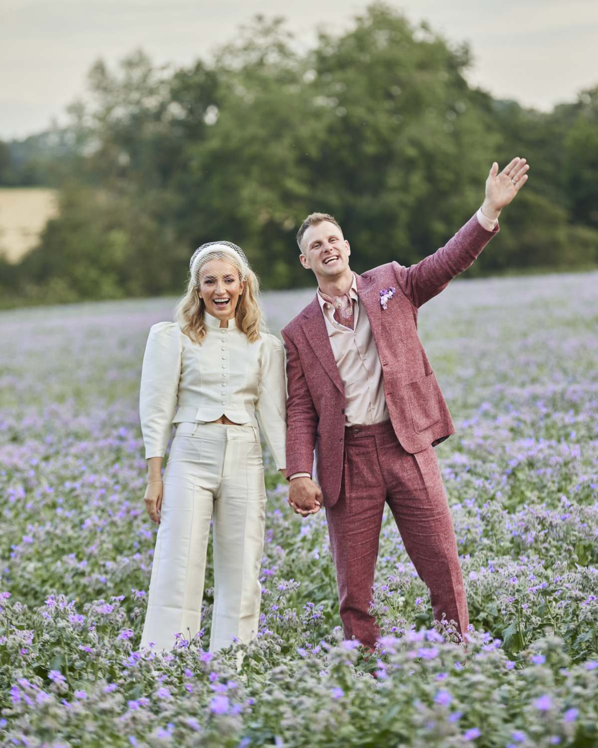 Alex in Emma Beaumont for her Ibiza & Disco Inspired Wedding at Wilderness Reserve 93 Bride and groom walking hand in hand through a field of lilac wildflowers, beaming with joy and waving — a relaxed and romantic wedding portrait full of personality and charm.