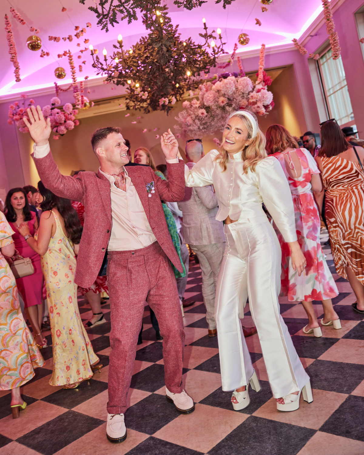 Alex in Emma Beaumont for her Ibiza & Disco Inspired Wedding at Wilderness Reserve 81 Bride and groom dancing joyfully during their wedding reception, with the bride in a chic ivory satin bridal jumpsuit and headband, and the groom in a relaxed pink suit — surrounded by guests beneath bold floral installations and vibrant pink lighting.