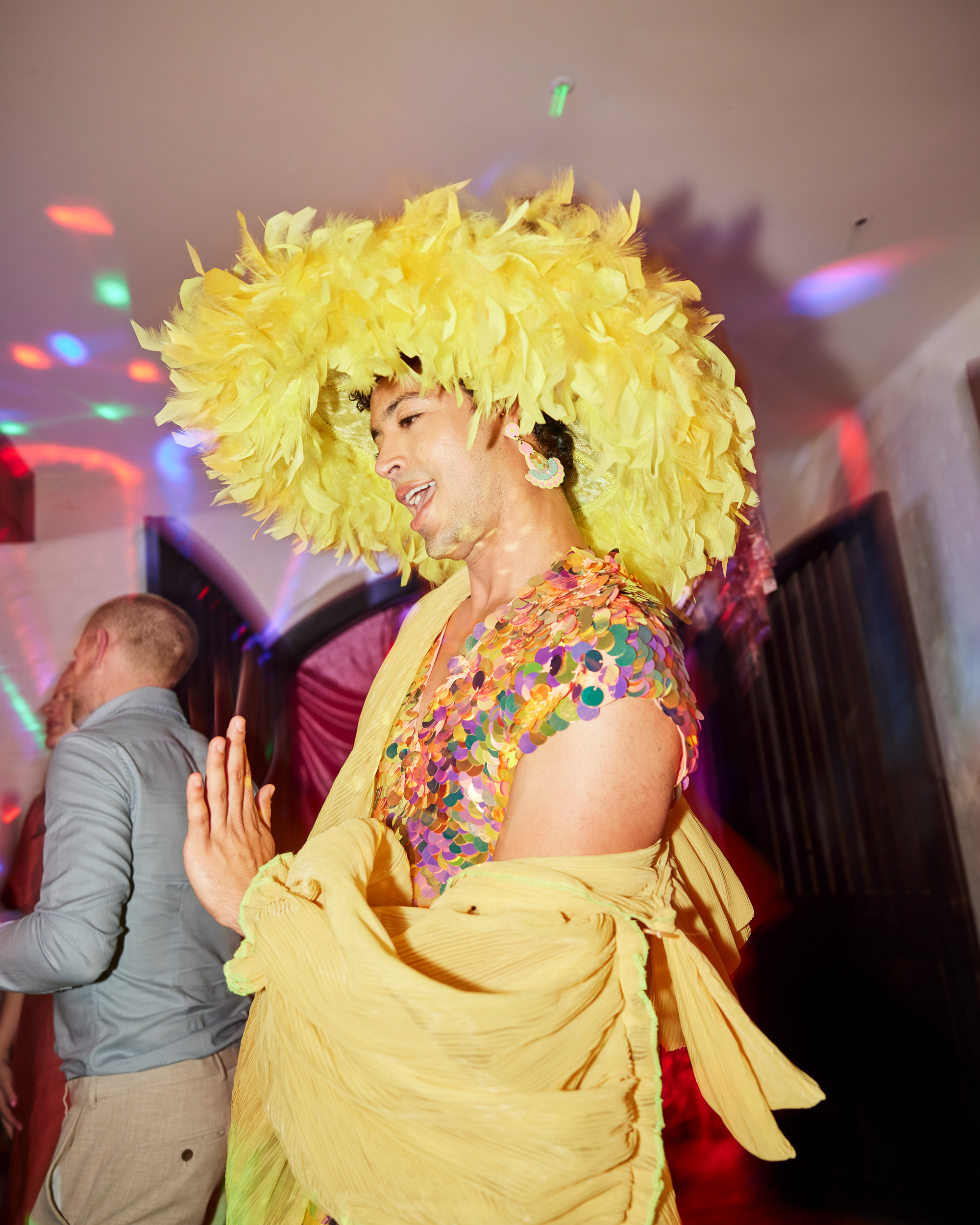 Wedding guest in a vibrant yellow feathered hat and sequin top lights up the dance floor — playful party style at a wedding full of bold colour and personality.