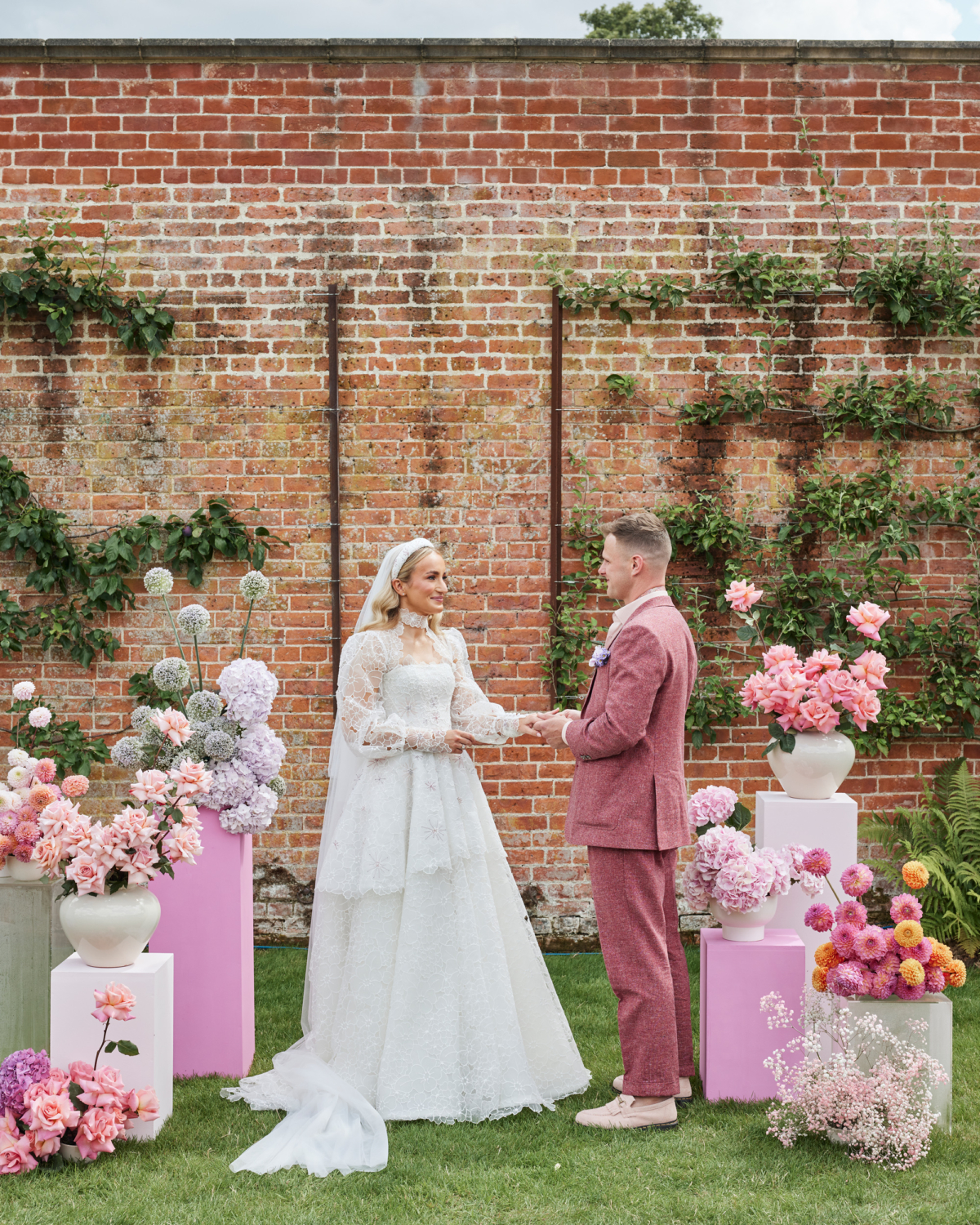 Alex in Emma Beaumont for her Ibiza & Disco Inspired Wedding at Wilderness Reserve 41 Bride and groom during their outdoor wedding reception at Wilderness Reserve. Bride wears a puff sleeve Emma Beaumont wedding dress, the groom wears a dark pink suit. They are surrounded by pink and white plinths with large colourful floral arrangements on top.