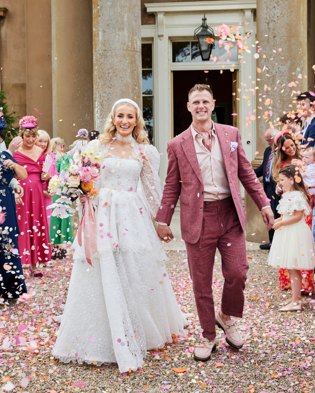Alex in Emma Beaumont for her Ibiza & Disco Inspired Wedding at Wilderness Reserve 49 Bride and groom being showered with pastel colour paper confetti outside Wilderness Reserve wedding venue. The groom wears a dark pink Beggars Run suit and the bride wears a bespoke Emma Beaumont dress and carries a pastel bouquet with pink ribbons. Joyous just married scene.