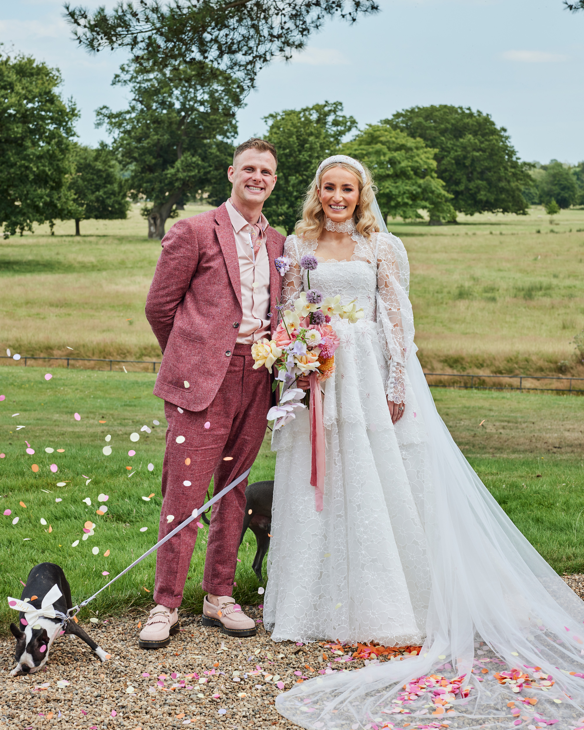 Alex in Emma Beaumont for her Ibiza & Disco Inspired Wedding at Wilderness Reserve 12 Bride wearing an Emma Beautmont dress with Lace Peplum with her groom in a dark pink suit by Beggars Run. The groom has hold of their dog on a lead. The dog wears a bow. Pastel colour paper confetti falls around them and the bride carries a large pastel colour bouquet. Wilderness Reserve wedding.
