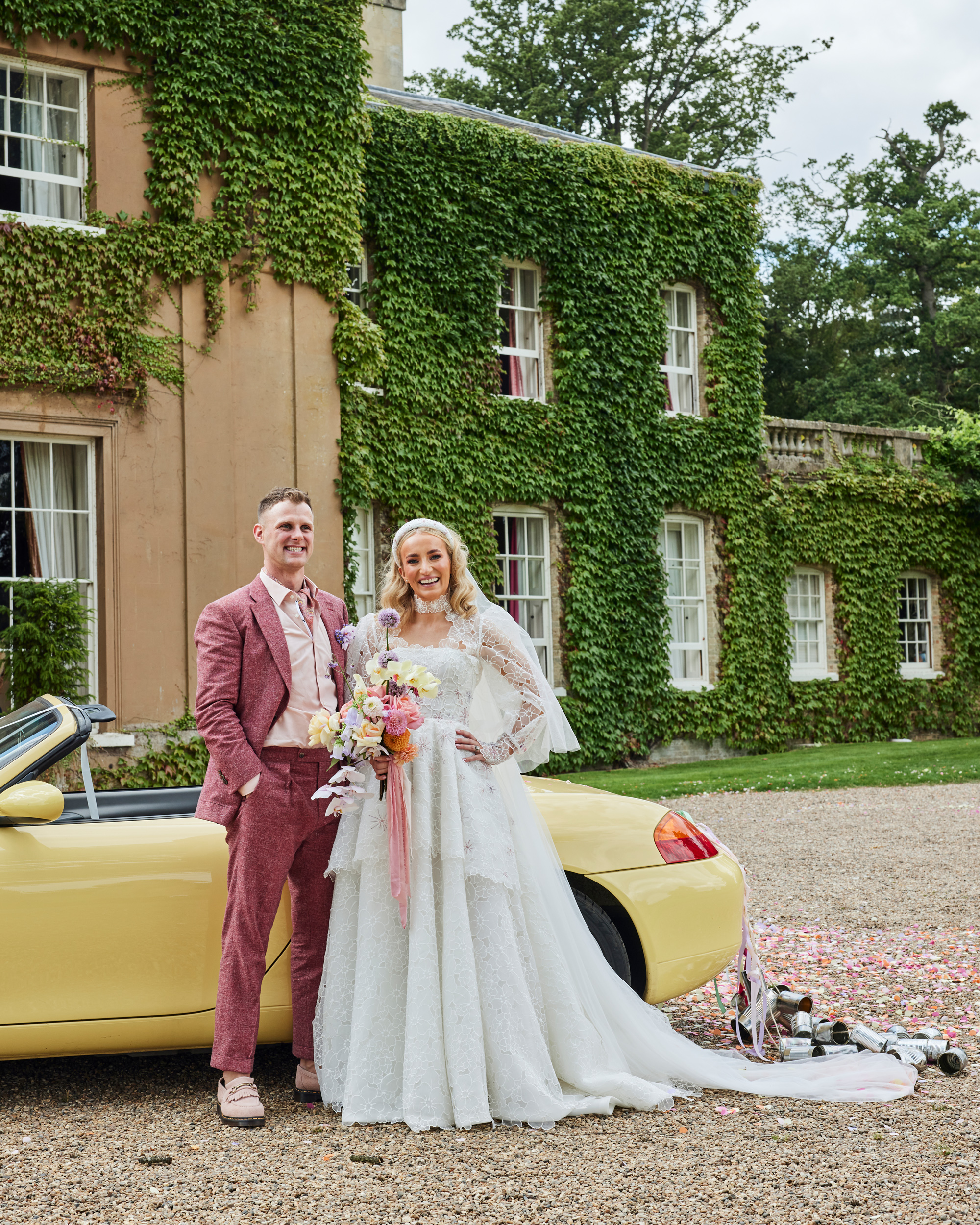 Alex in Emma Beaumont for her Ibiza & Disco Inspired Wedding at Wilderness Reserve 48 Bride wearing a puff sleeve Emma Beaumont wedding dress and groom wearing a dark pink suit standing next to a yellow sports car with traditional tin cans and ribbons tied to the back. Wilderness Reserve wedding.