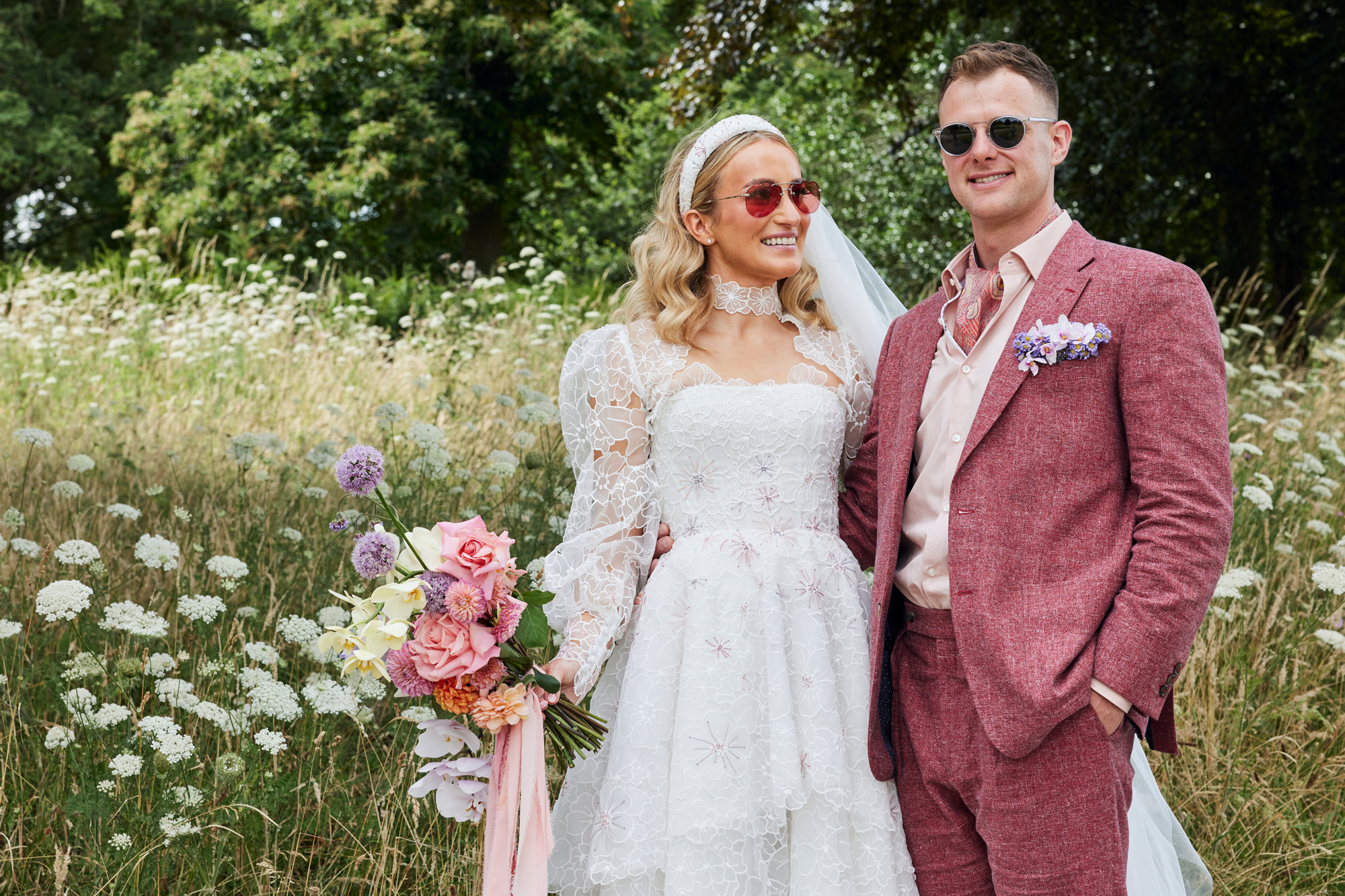 Bride in sunglasses and an Emma Beaumont ATelier gown. She carries a pastel & pink bouquet. Groom wears a pink suit by Beggars Run.