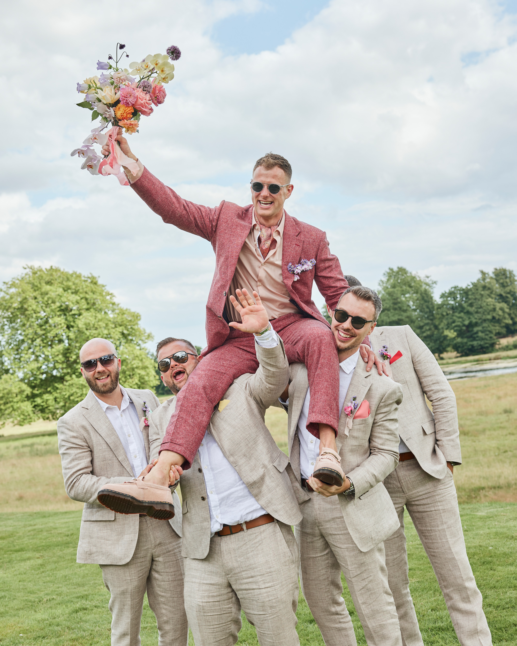 Alex in Emma Beaumont for her Ibiza & Disco Inspired Wedding at Wilderness Reserve 92 Groom lifted on the shoulders of his groomsmen, holding the bridal bouquet in triumph — a playful and lighthearted portrait in relaxed linen suits and summer wedding style.