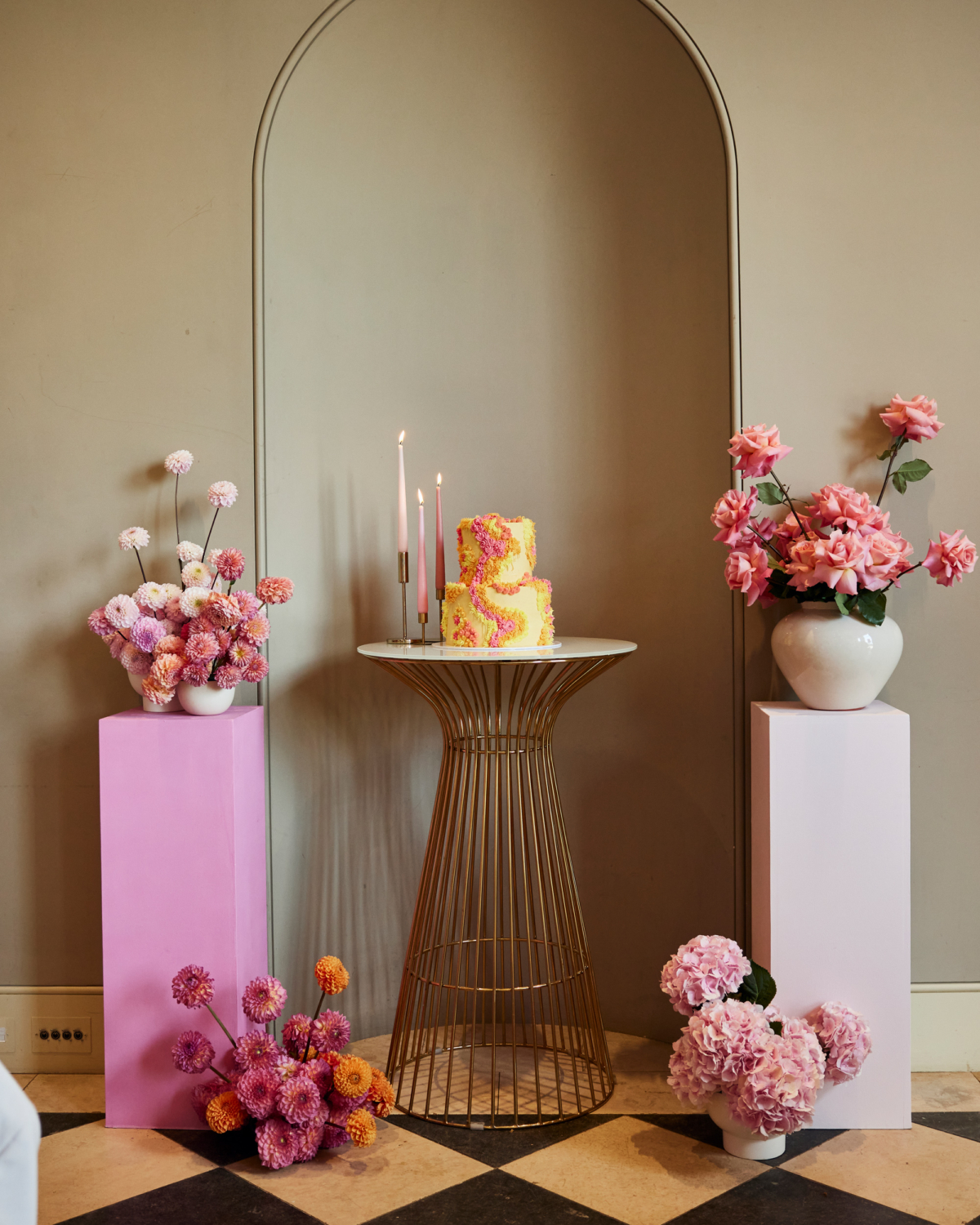 Alex in Emma Beaumont for her Ibiza & Disco Inspired Wedding at Wilderness Reserve 72 2 tier yellow wedding cake with pink and yellow ruffled icing, standing on a gold bar table with plinths either side that have colourful flower arrangements in bowls.