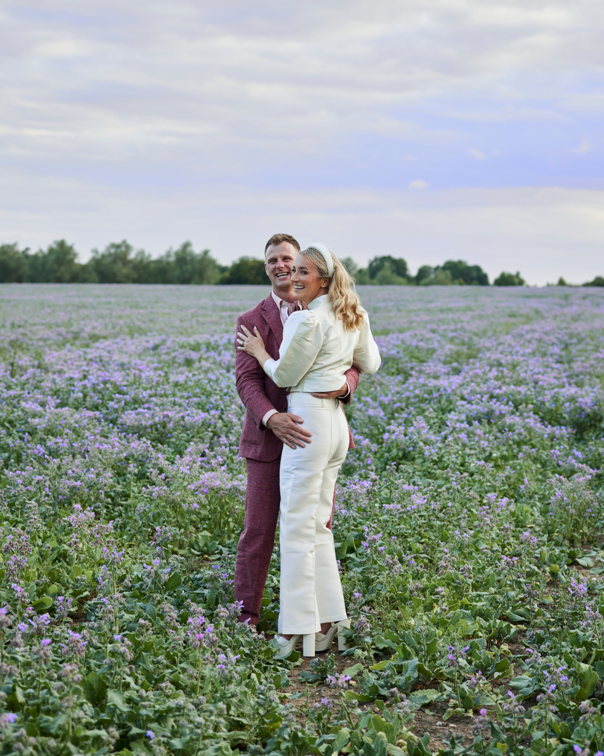 Alex in Emma Beaumont for her Ibiza & Disco Inspired Wedding at Wilderness Reserve 94 Newlyweds embracing in a meadow of soft purple wildflowers under a pastel sky — a romantic and carefree moment between bride and groom in modern wedding day attire.