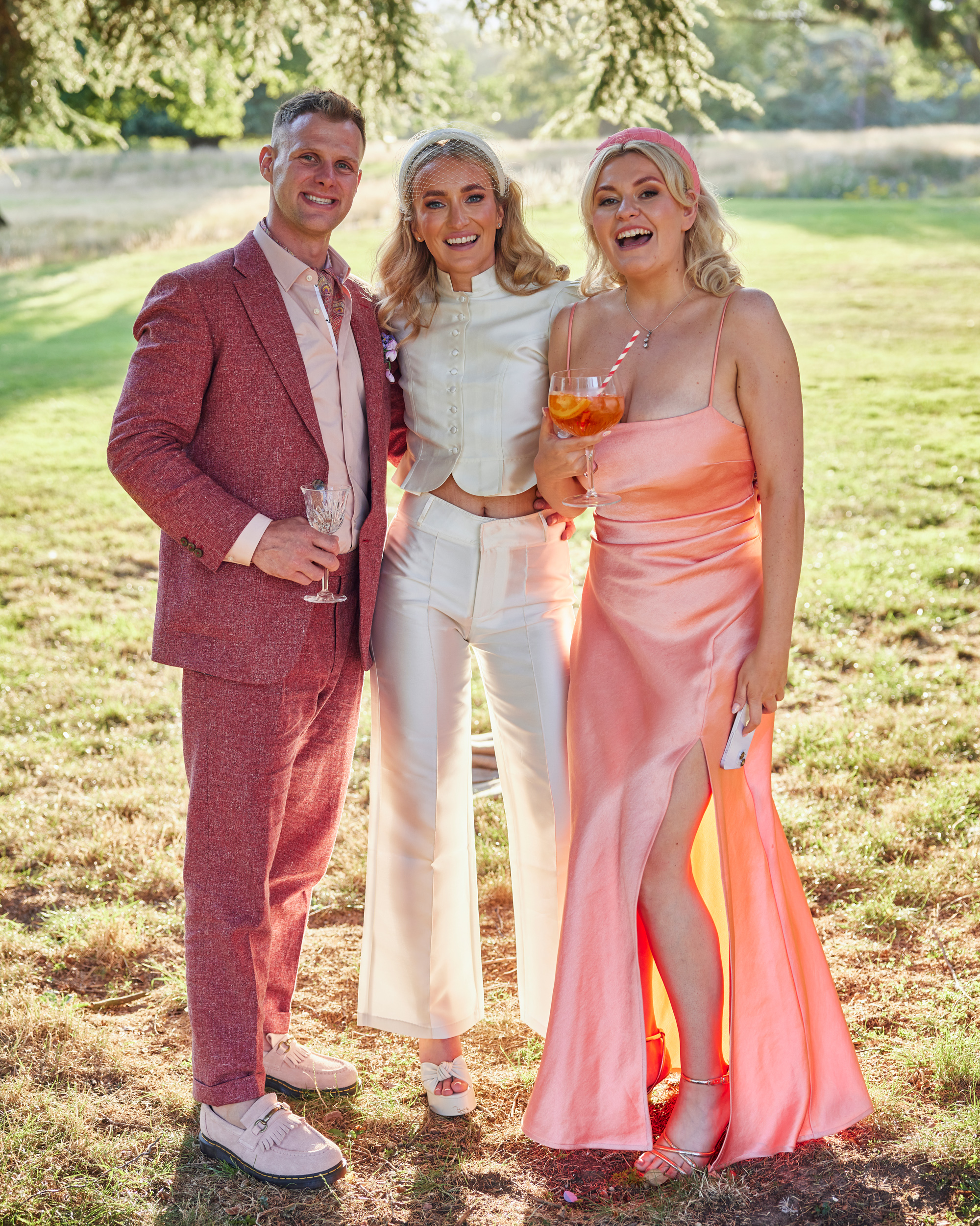 Alex in Emma Beaumont for her Ibiza & Disco Inspired Wedding at Wilderness Reserve 83 Bride and groom pose with a bridesmaid in the sunshine, all holding cocktails and dressed in stylish modern wedding attire — featuring the bride in a chic ivory jumpsuit and the groom in a relaxed dusky pink suit and the bridesmaid in a peach dress with a thigh slit.