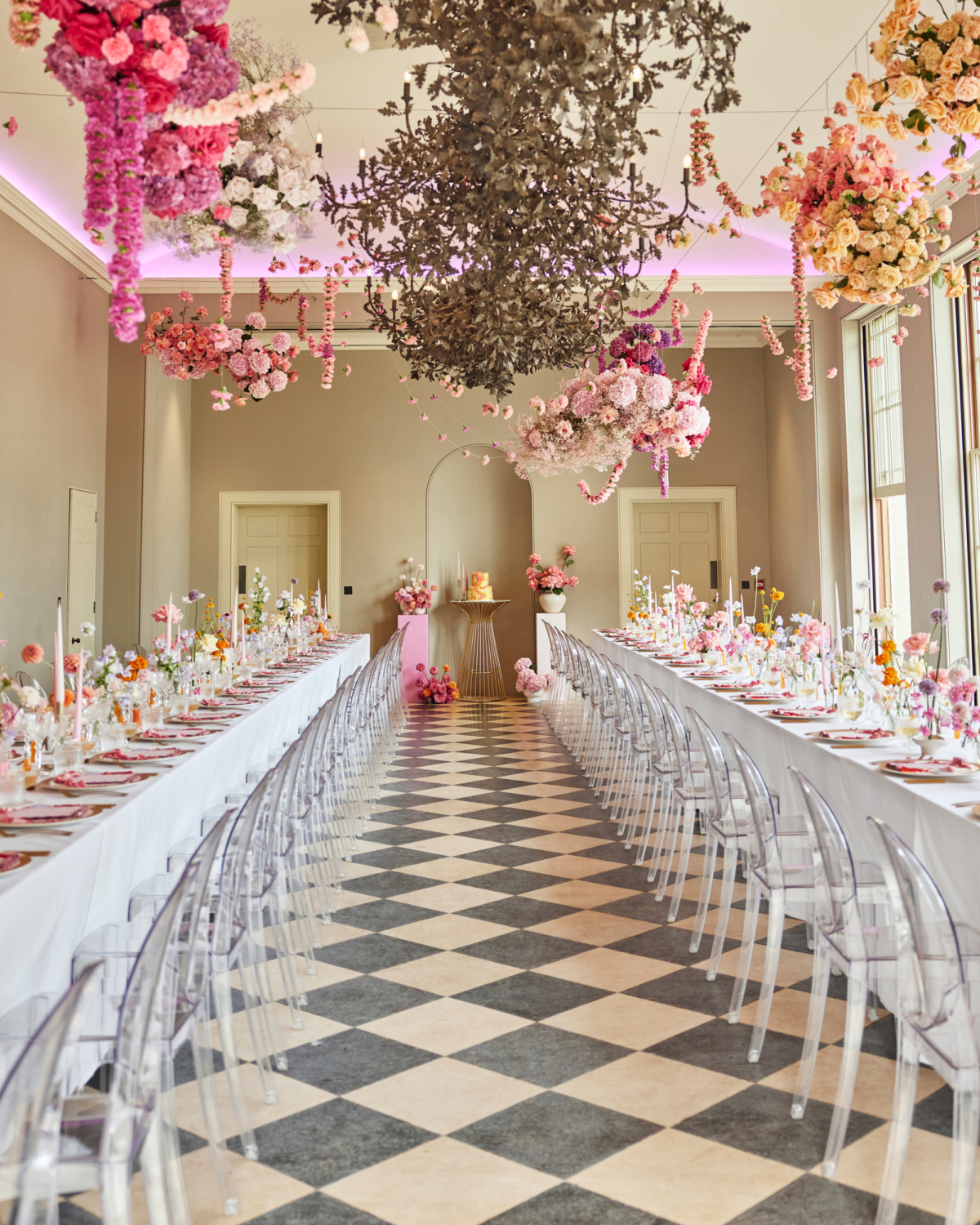 Alex in Emma Beaumont for her Ibiza & Disco Inspired Wedding at Wilderness Reserve 55 Wilderness Reserve wedding reception room. Chequerboard floor with 2 long rows of dining tables and ghost chairs. The tables have white linen and are covered in pink table decor including pink napkins, wedding stationery, taper candles and flowers. Above the diners, a riot of pastel colour flowers are suspended from the ceiling.