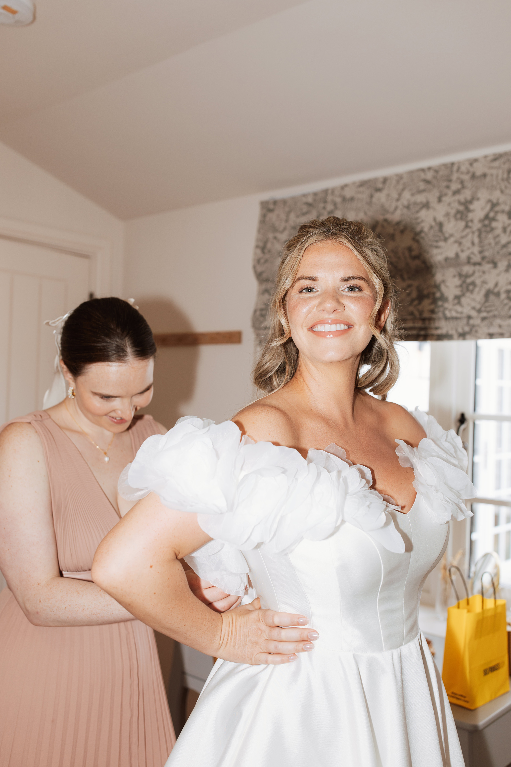 Laura & Alex's Colourful, Black Tie Wedding at Woodmancote Place 53 Bridesmaid in a pale pink dress buttoning up a Royaldi wedding dress as the bride smiles into the camera.