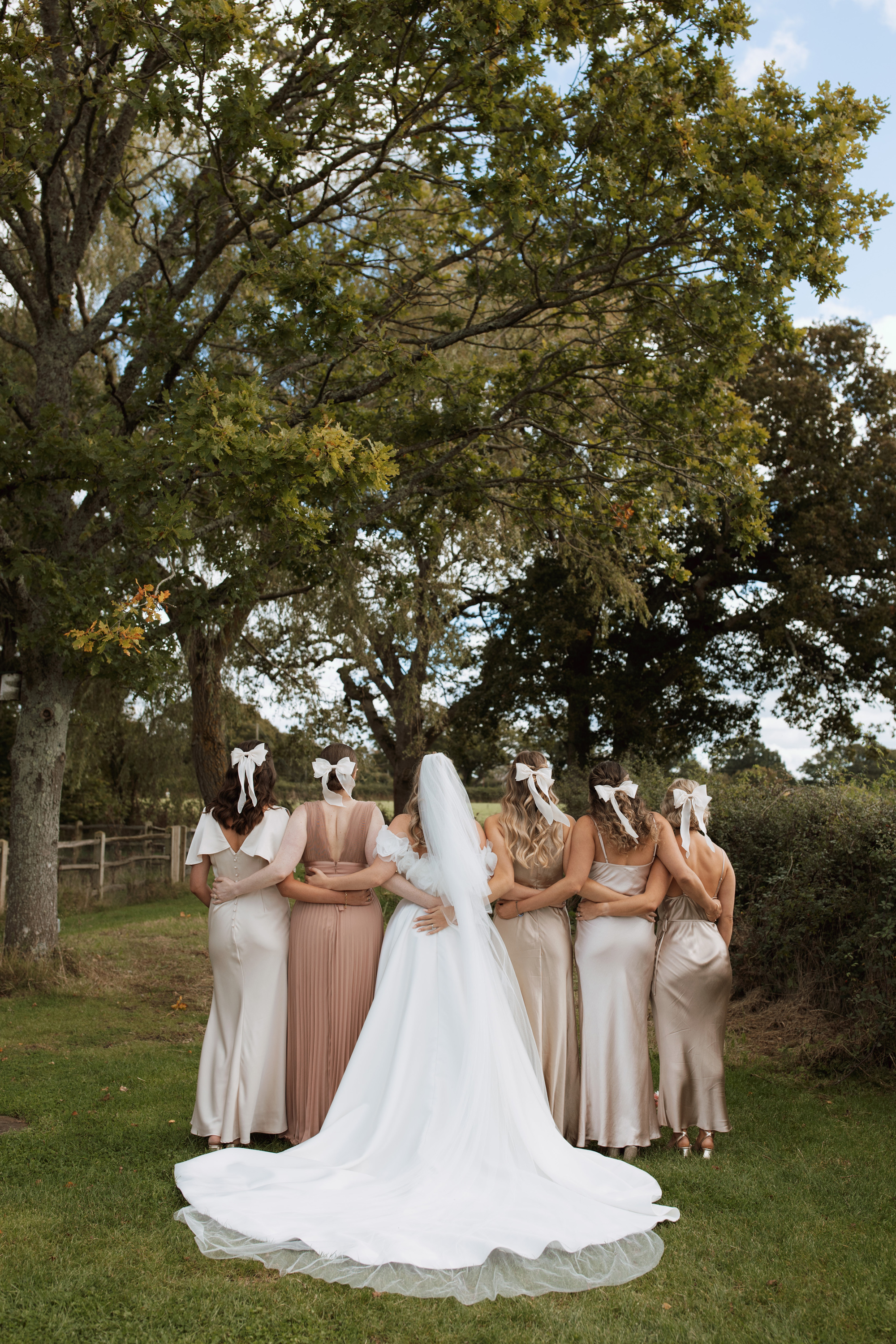 Laura & Alex's Colourful, Black Tie Wedding at Woodmancote Place 56 Photograph from behind a bride and her bridesmaids as they look in the opposite direction. The bridesmaids all wear bows in their hair and the bridesmaids wears a Ronaldi dress with a long veil.