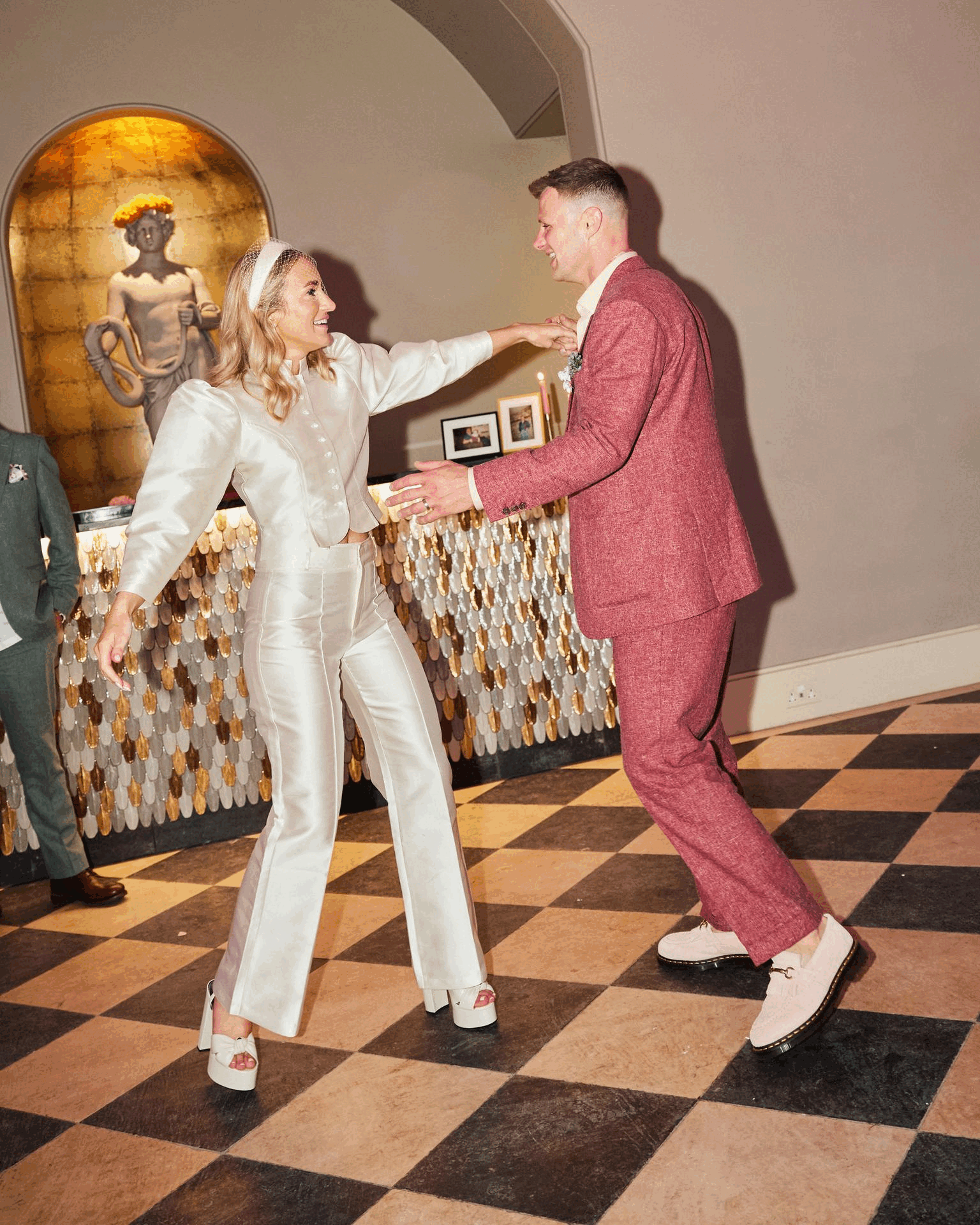 Newlyweds share their first dance, with the bride in a sleek ivory satin jumpsuit and the groom in a relaxed pink suit — a joyful moment full of fun and connection.