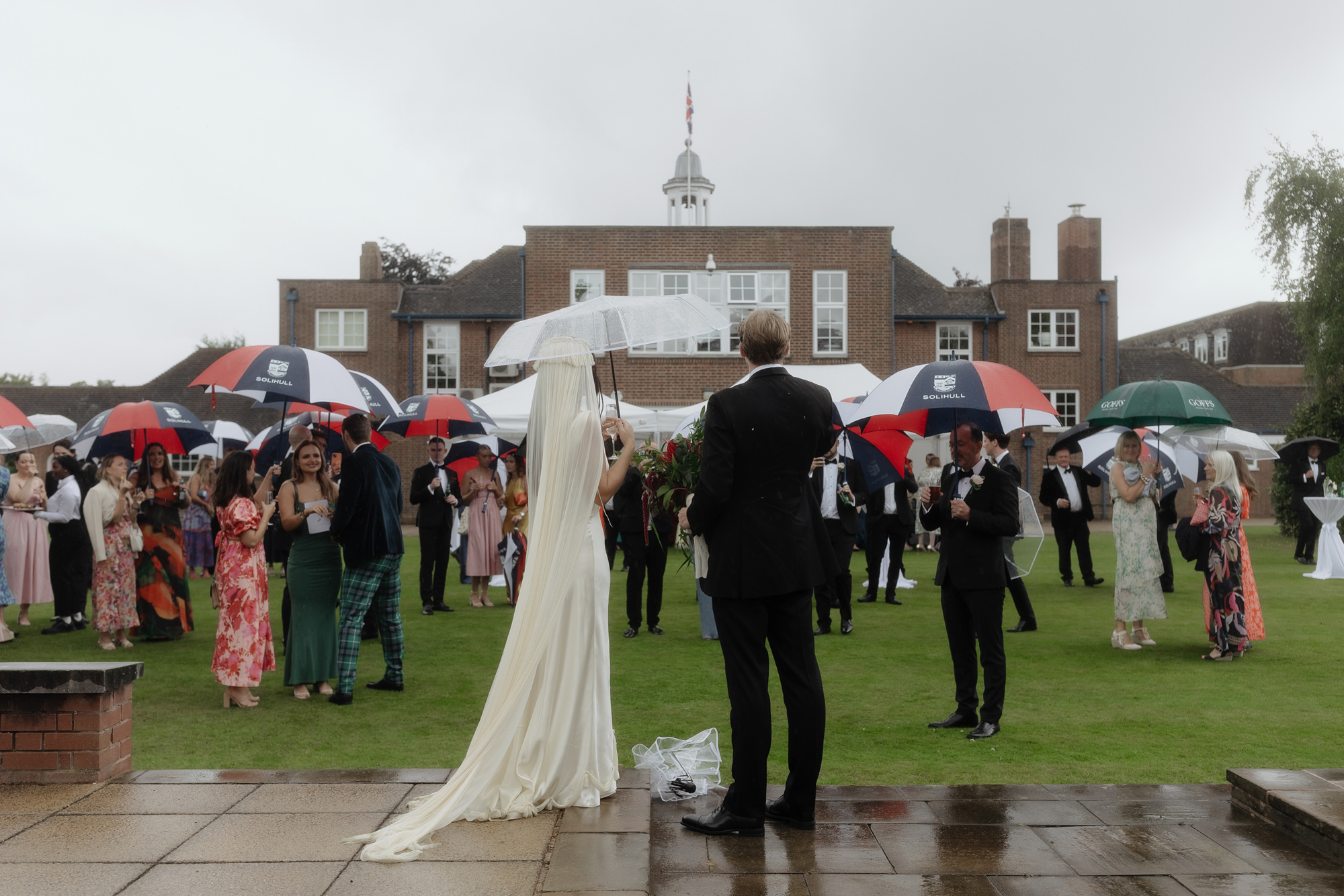 Photograph of the back of a bride and groom standing in the rain as they look on to their wedding guests carrying umbrellas.