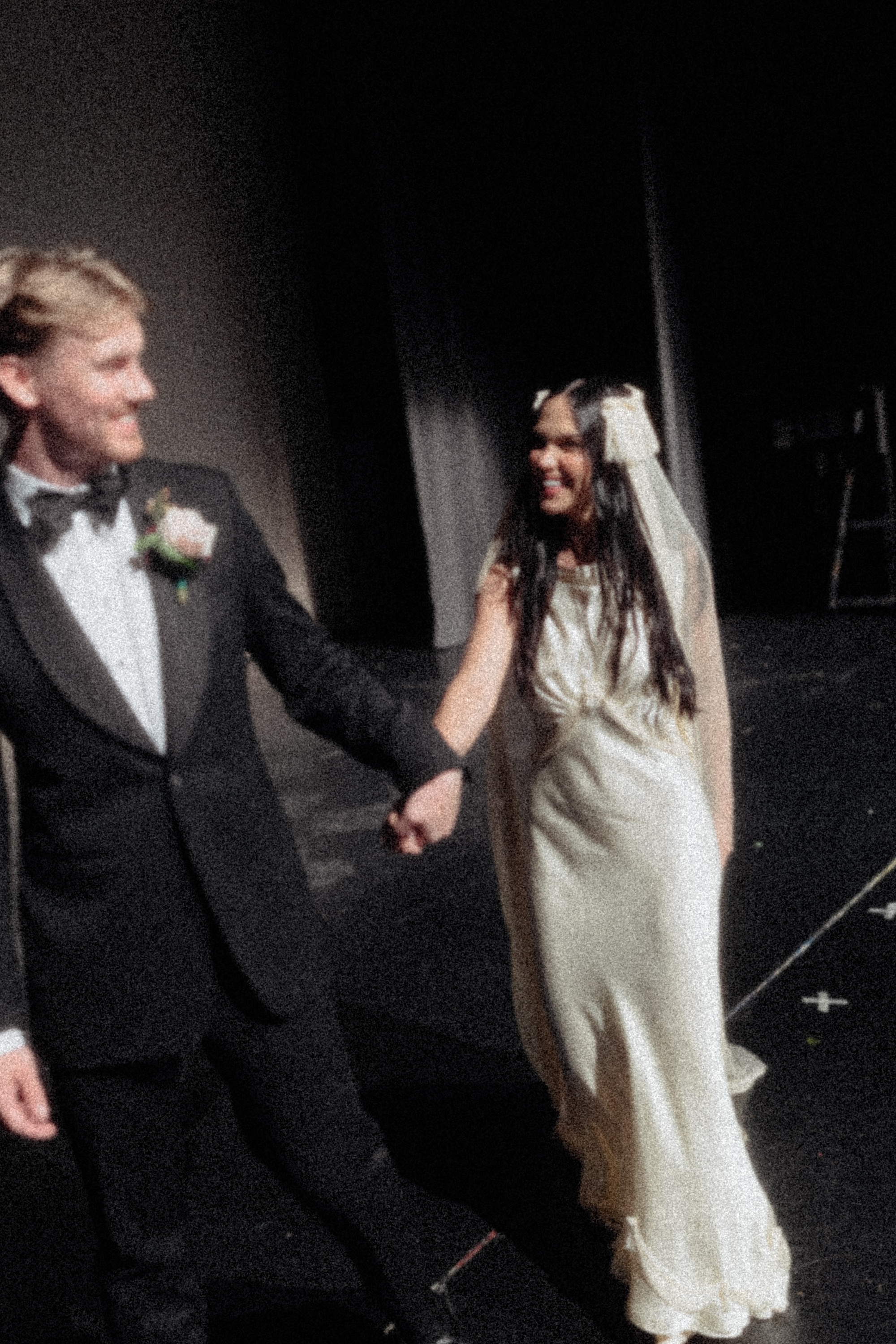 Grainy film shot of a bride in a Paula Nanda wedding dress and fringed veil, holding the hand of her husband in black tie with a pink rose buttonhole
