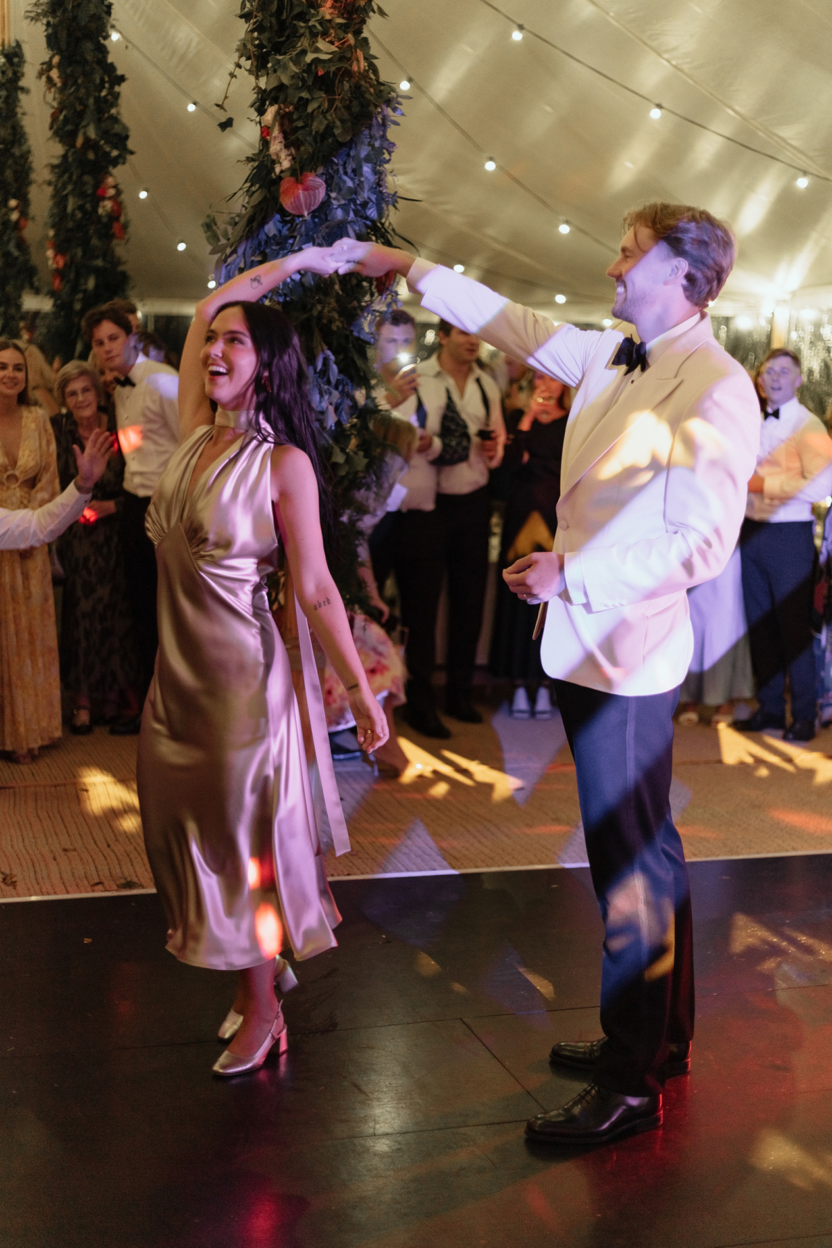Groom twirling the bride around on the dance floor. He wears a white tuxedo and she wears a silver 