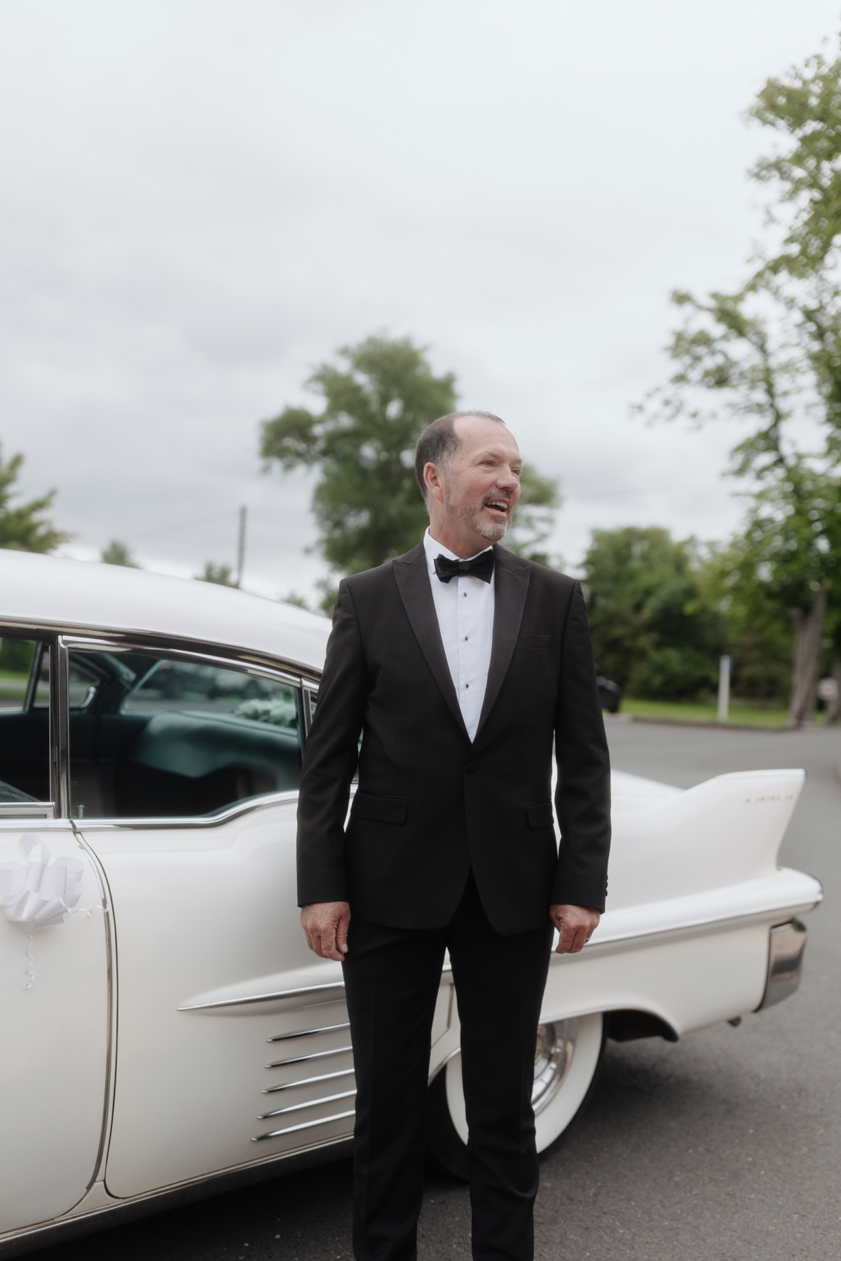 Father of the bride in black tie standing next to a vintage white wedding car.