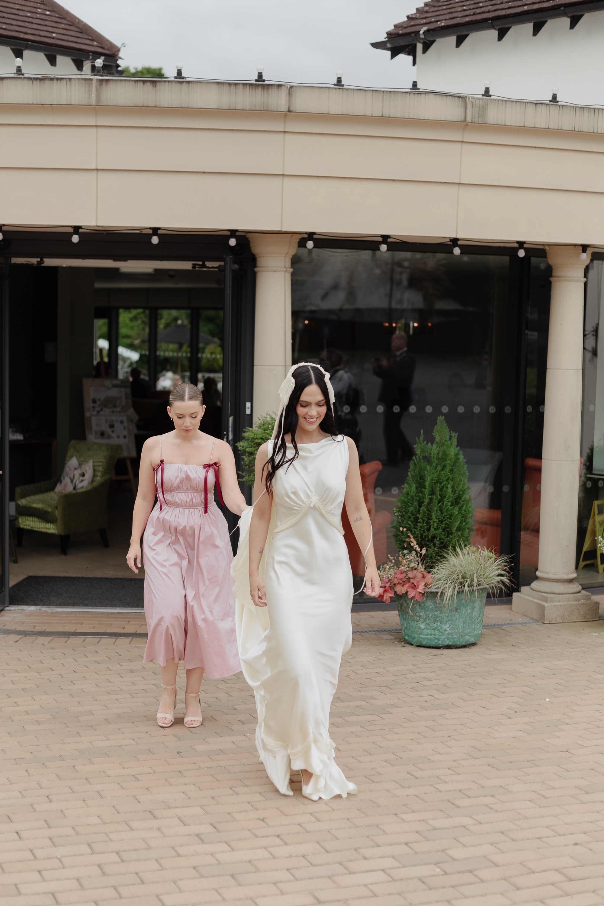 Bride stepping outside in her vintage inspired Paula Nanda wedding dress and fringed tasseled wedding veil. Behind her a bridesmaid in a pink Cos dress holds her dress train.