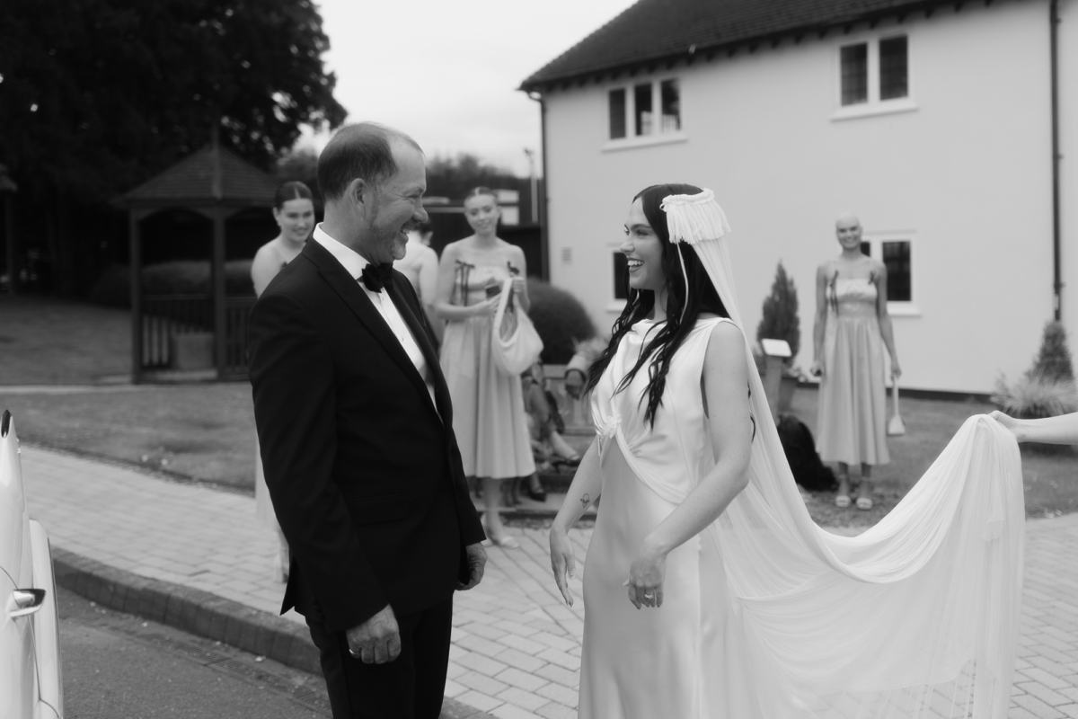 Bride in a Paula Nanda dress and fringed tasseled wedding veil meeting her father who is wearing black tie.