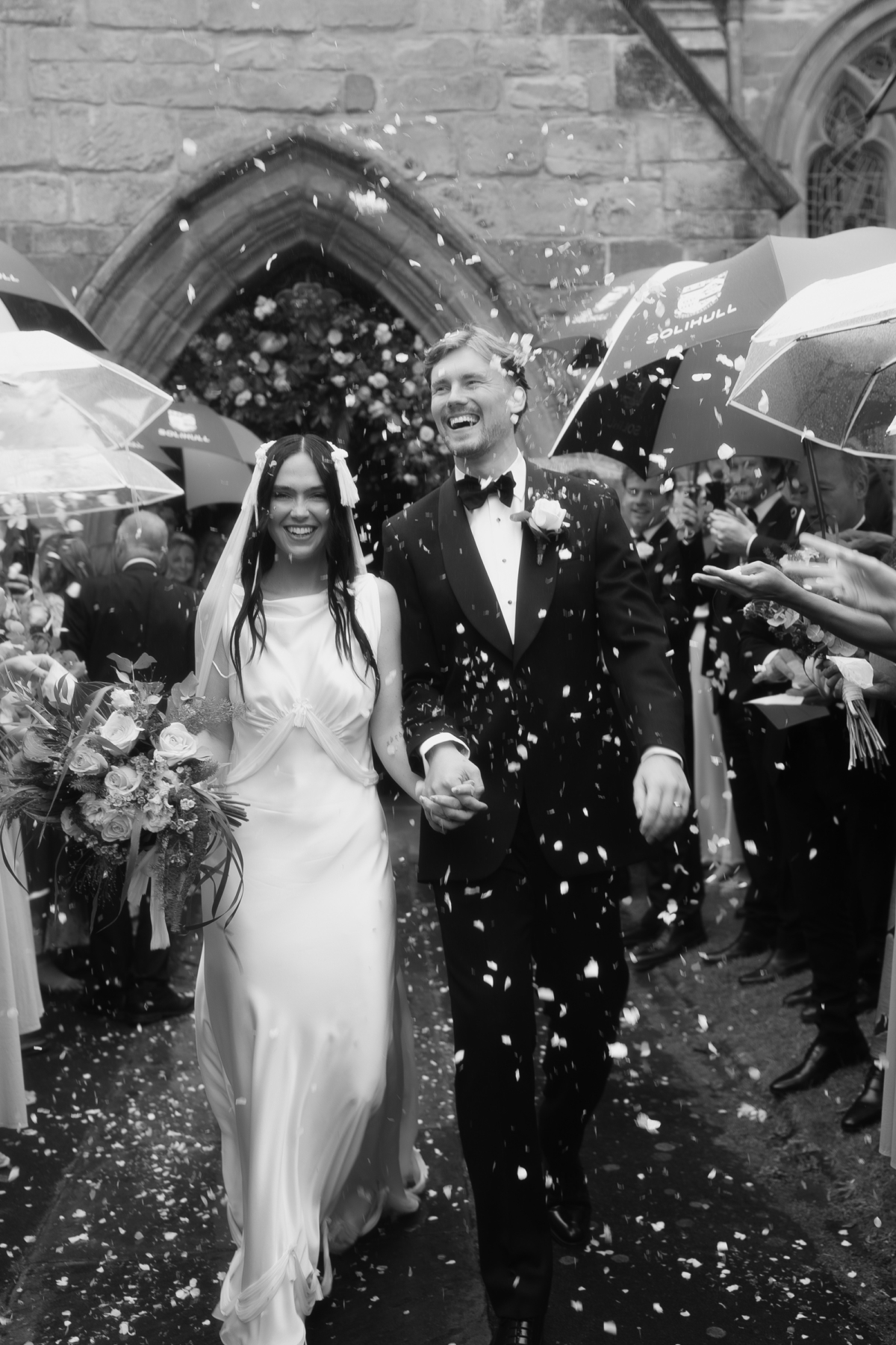 Confetti shot of bride and groom leaving church. it is raining and the guests carry umbrellas. The bride wears a Paula Nanda wedding dress & fringed veil.