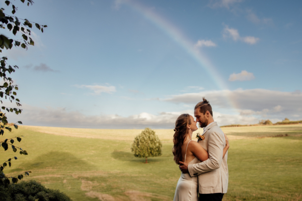 A bride and groom embrace is front of a rainbow at their wedding in Devon.