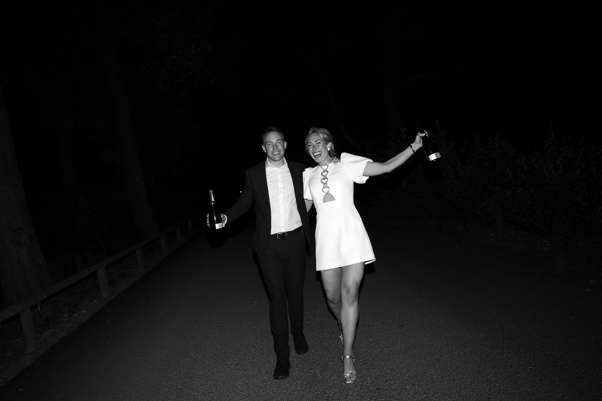 Black and white photograph of bride and groom each holding a champagne bottle on their wedding night. She wears a Rebecca Vallance short white dress.