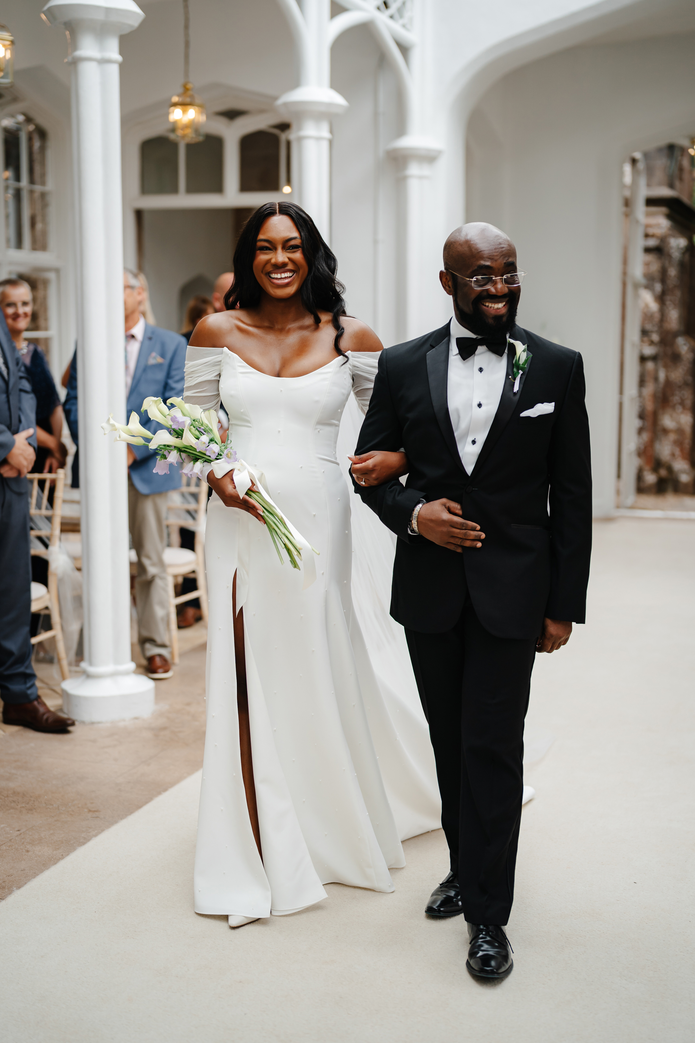 Black bride arm in arm wiht her father walking down the aisle at St Audries Park in Somerset. She wears Bronte by Sarah Seven and carries a bouquet of calla lillies. Jodie Hurd Photography.