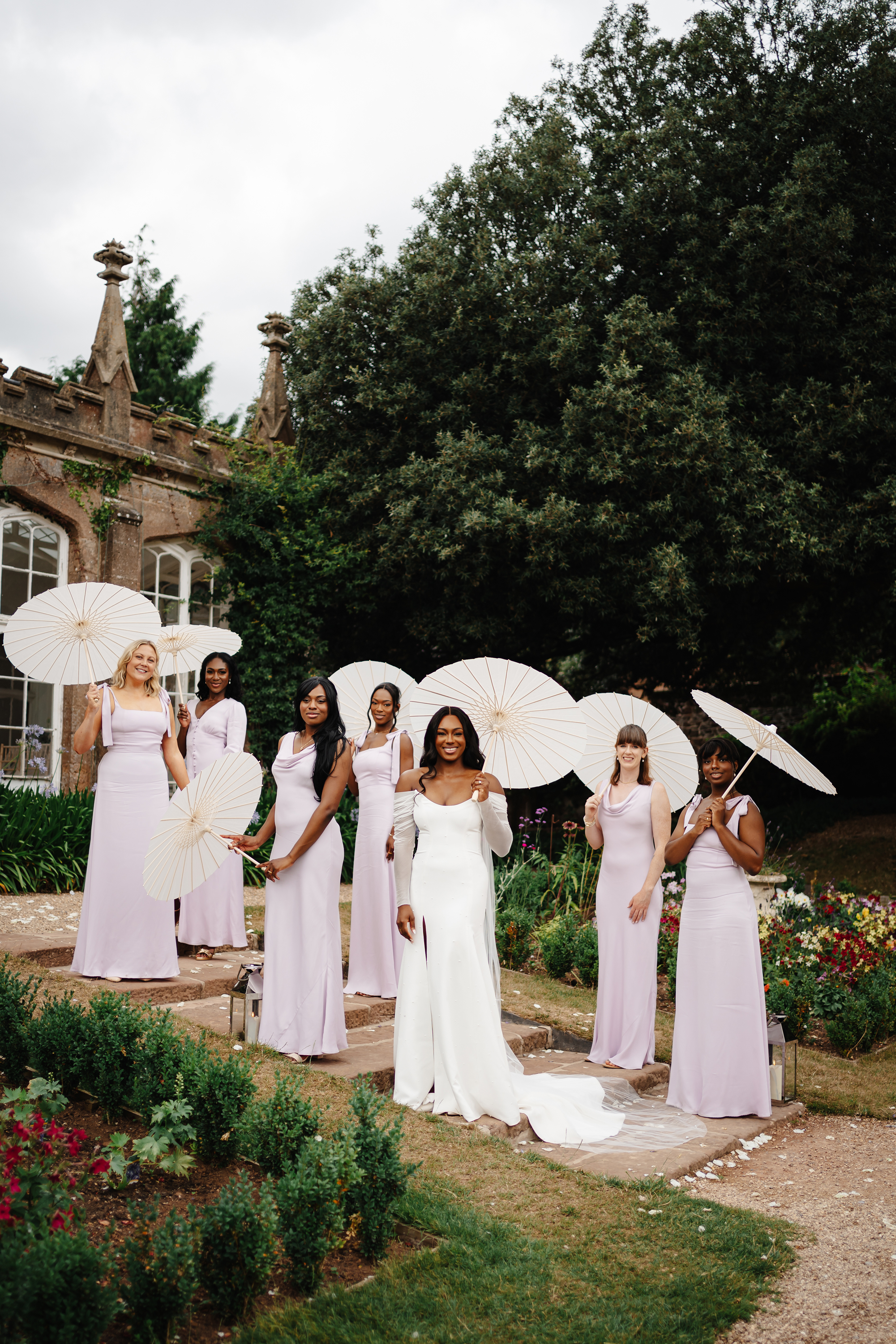 Radiant and elegant Black bride with her bridesmaids wearing lilac dresses by Maids To Measure.  They are carrying parasols and standing in. the gardens of St Audries Park wedding venue in Somerset.