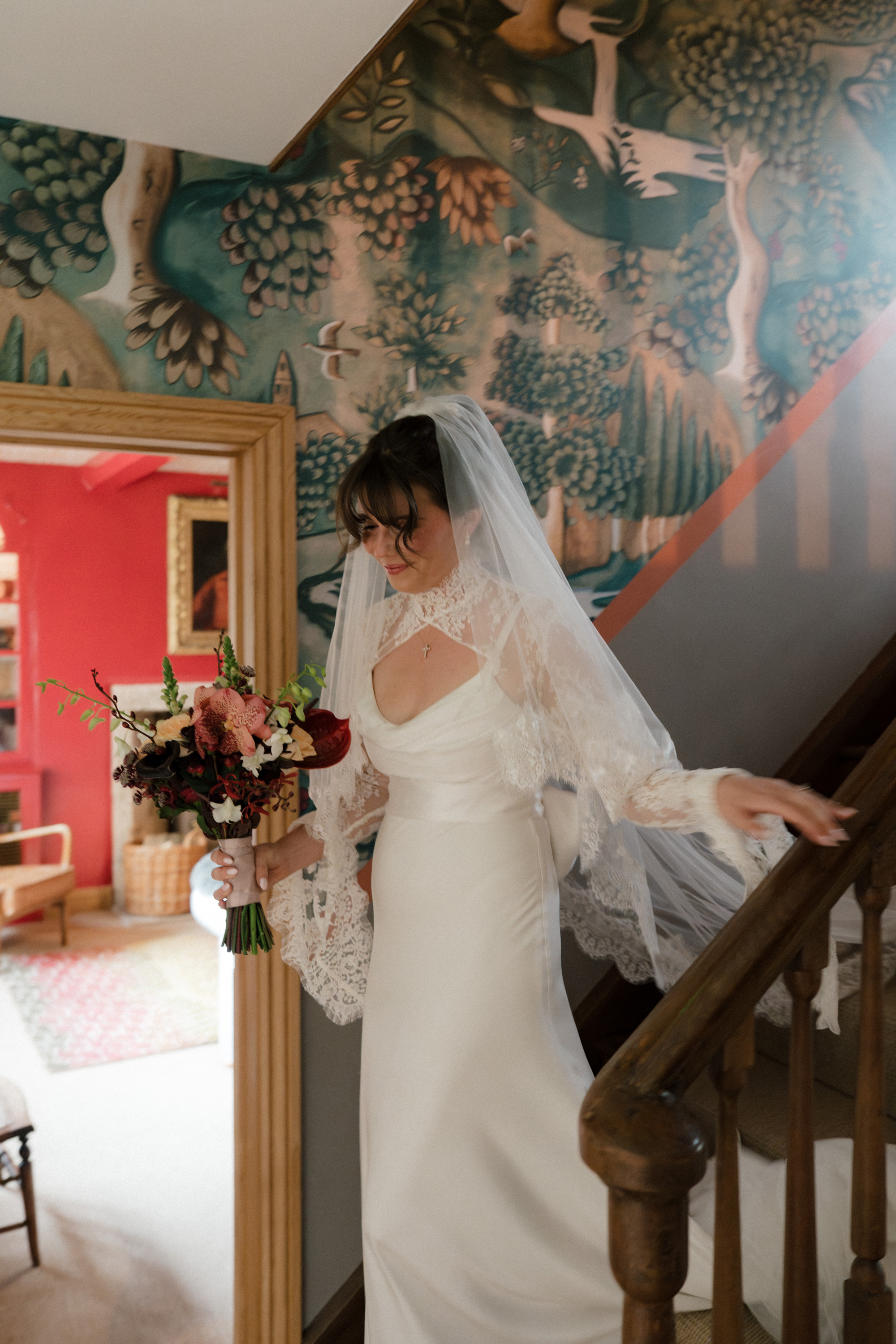 Bride wearing Saint Bridal Couture and holding a small bouquet of red and pink flowers, walking down the stairs at Gulliver's Hall, a Unique Home Stays wedding venue in The Cotswolds.