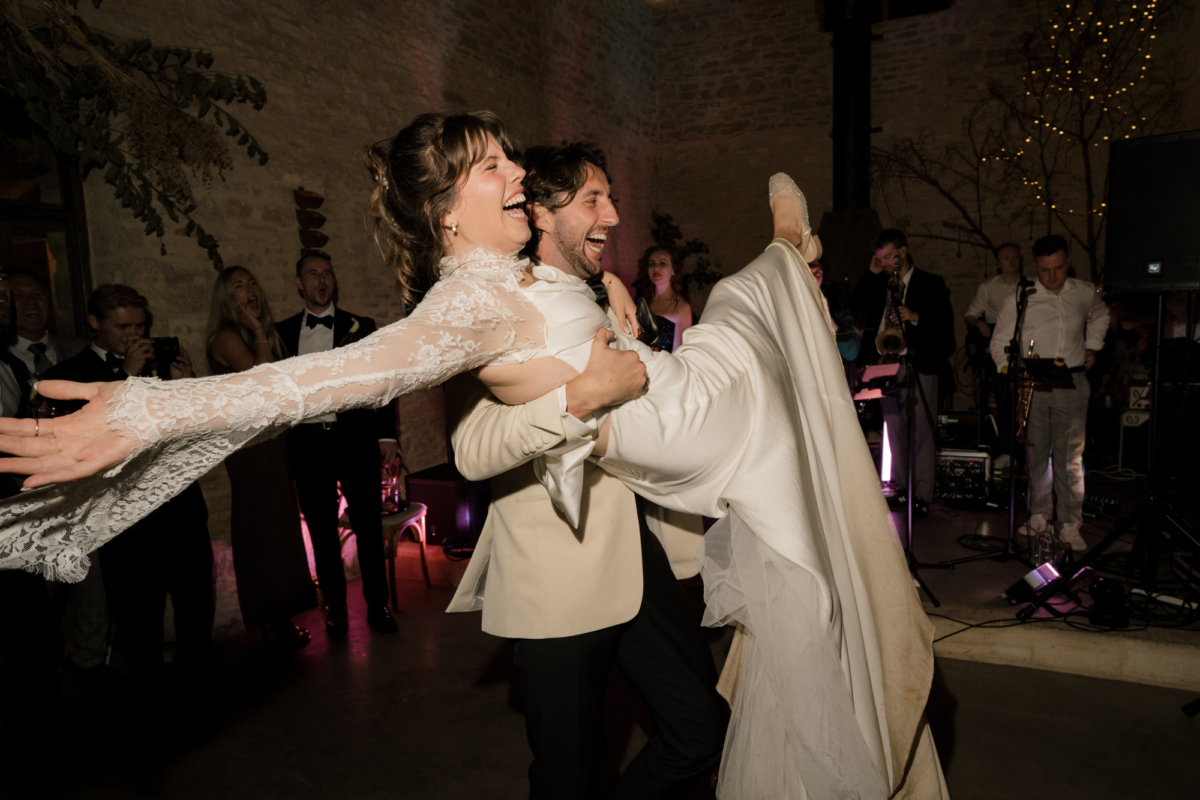 Wedding at Gulliver's Hall, a Unique Home Stays wedding venue in The Cotswolds. The groom wears a white tuxedo and is lifting the bride in her Saint Bride Couture dress on the dance floor.