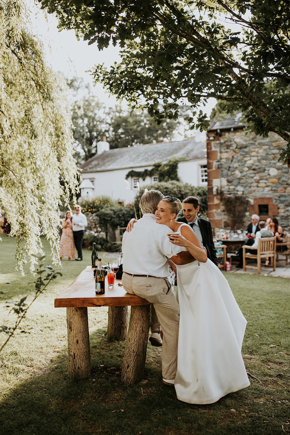 Bride leaning into her dad for a hug on her wedding day. She stands outdoors and is hlding a glass of champagne in one hand whilst her dad rests on a wooden table at Low Hall The Lakes rustic English country wedding venue in The Lake District.