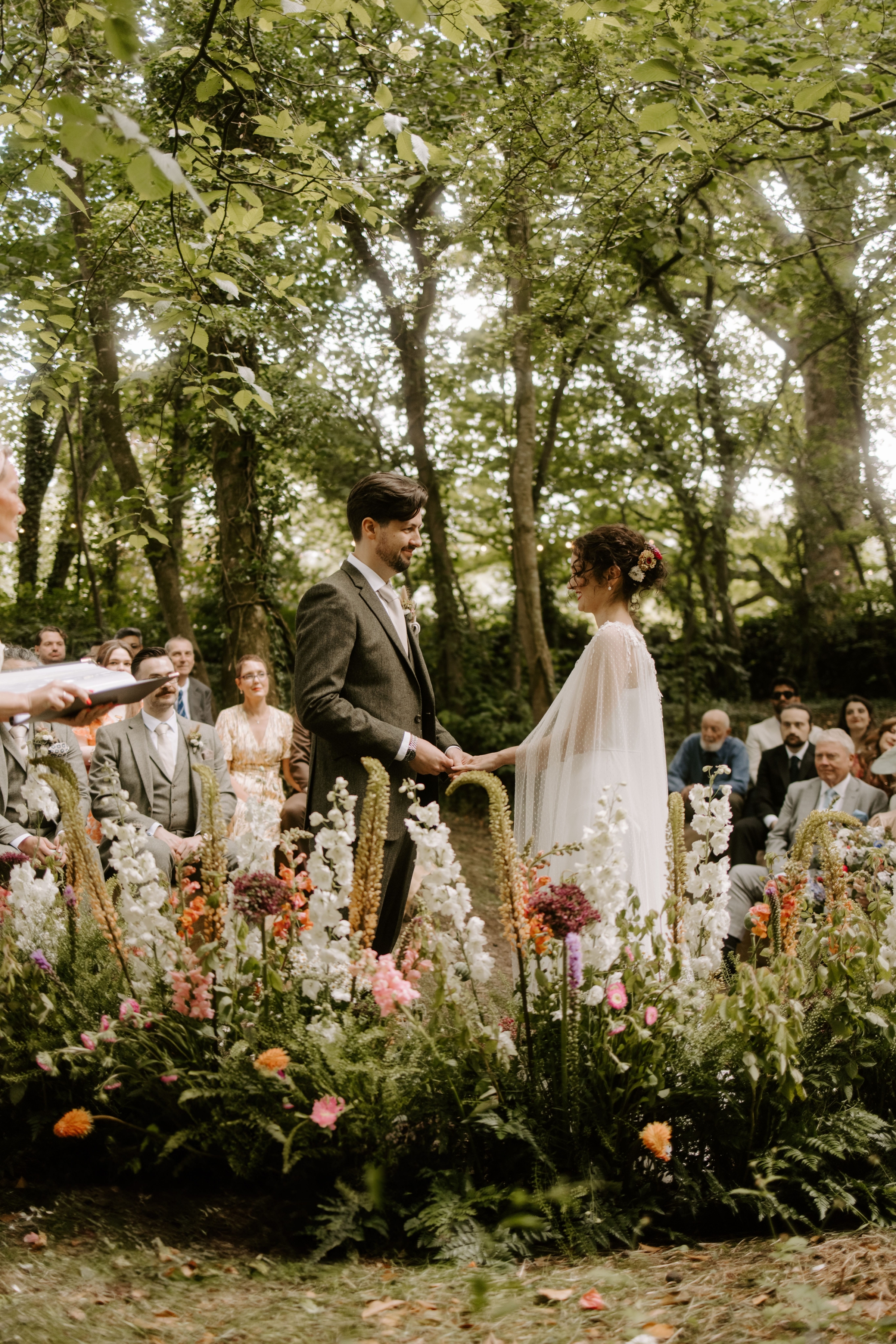  Bride and groom holding hands and looking into each others eyes during a magical outdoor wedding ceremony at Low Hall The Lakes in The Lake District. They are surrounded by trees, sunlight, wedding guests and a meadow flower installation.