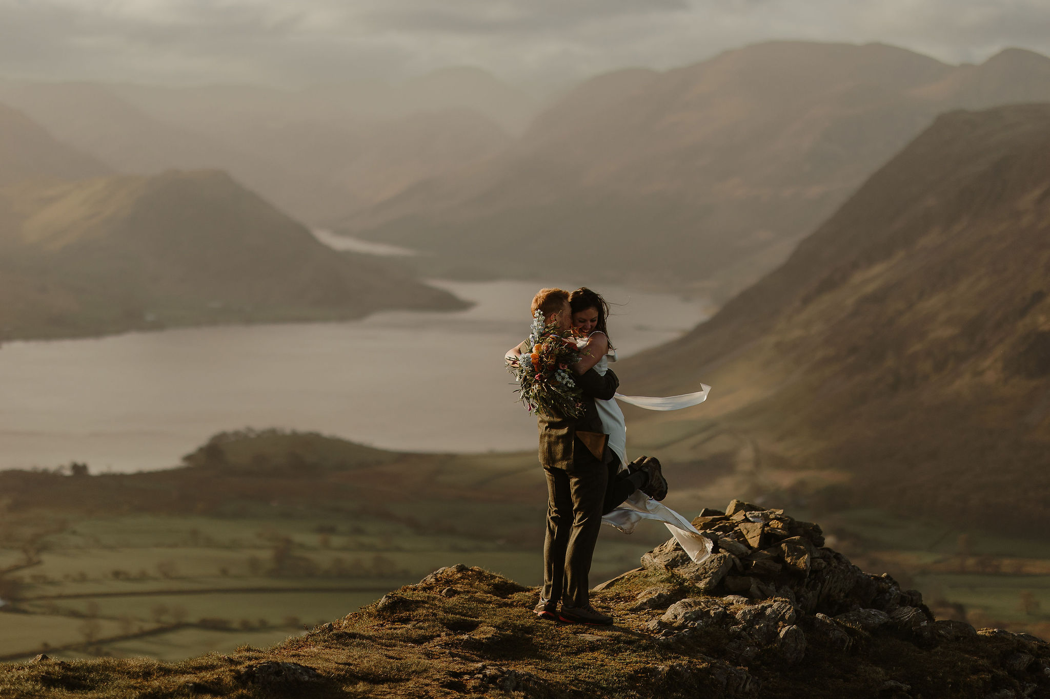 Beautiful outdoor landscape shot of a bride and groom hugging, the bride's bouquet ribbon captured in the breeze, behind them are epic lakes and mountains. Wedding at Low Hall The Lakes rustic English country wedding venue in The Lake District.
