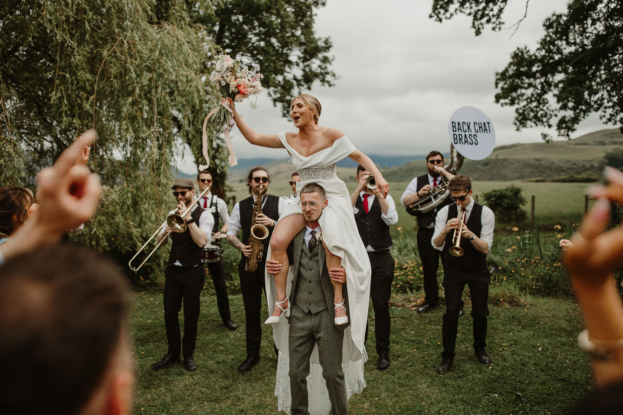 Groom carrying his bride on his shoulders. Behind them they are serenaded by the Back Chat Brass band - a wandering brass band who perform at weddings. Wedding at Low Hall The Lakes rustic English country wedding venue in The Lake District.