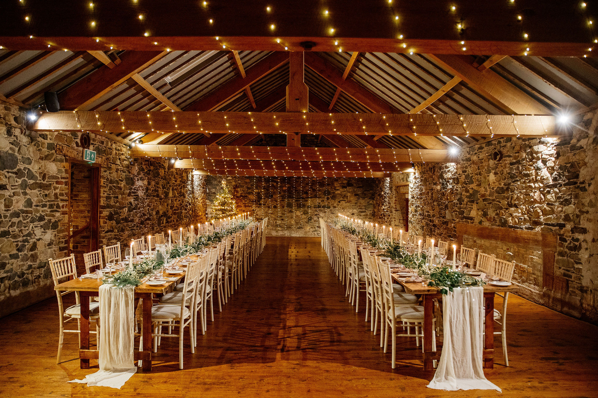 The Loveable Barn at Low Hall The Lakes, a pretty Lake District Wedding Venue. The barn is decorated with fairy lights and trestle tables that are lined with taper candles, linen runners and greenery.