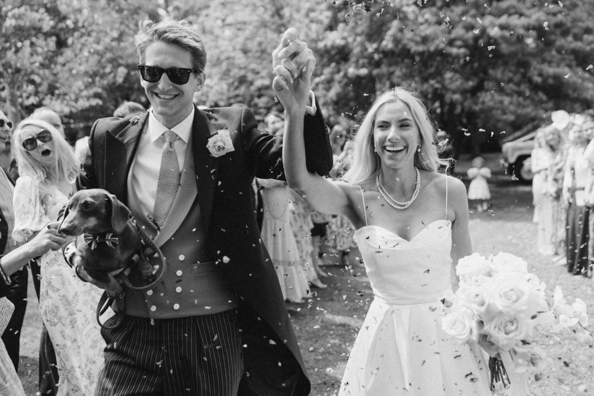 A Beautiful Summer Marquee Wedding at Home in the English Countryside 43 Black and white photograph of bride and groom exiting their outdoor ceremony to a shower of confetti. The bride wears a simple Vera Wang dress and carries a bouquet of tall stepped white roses.