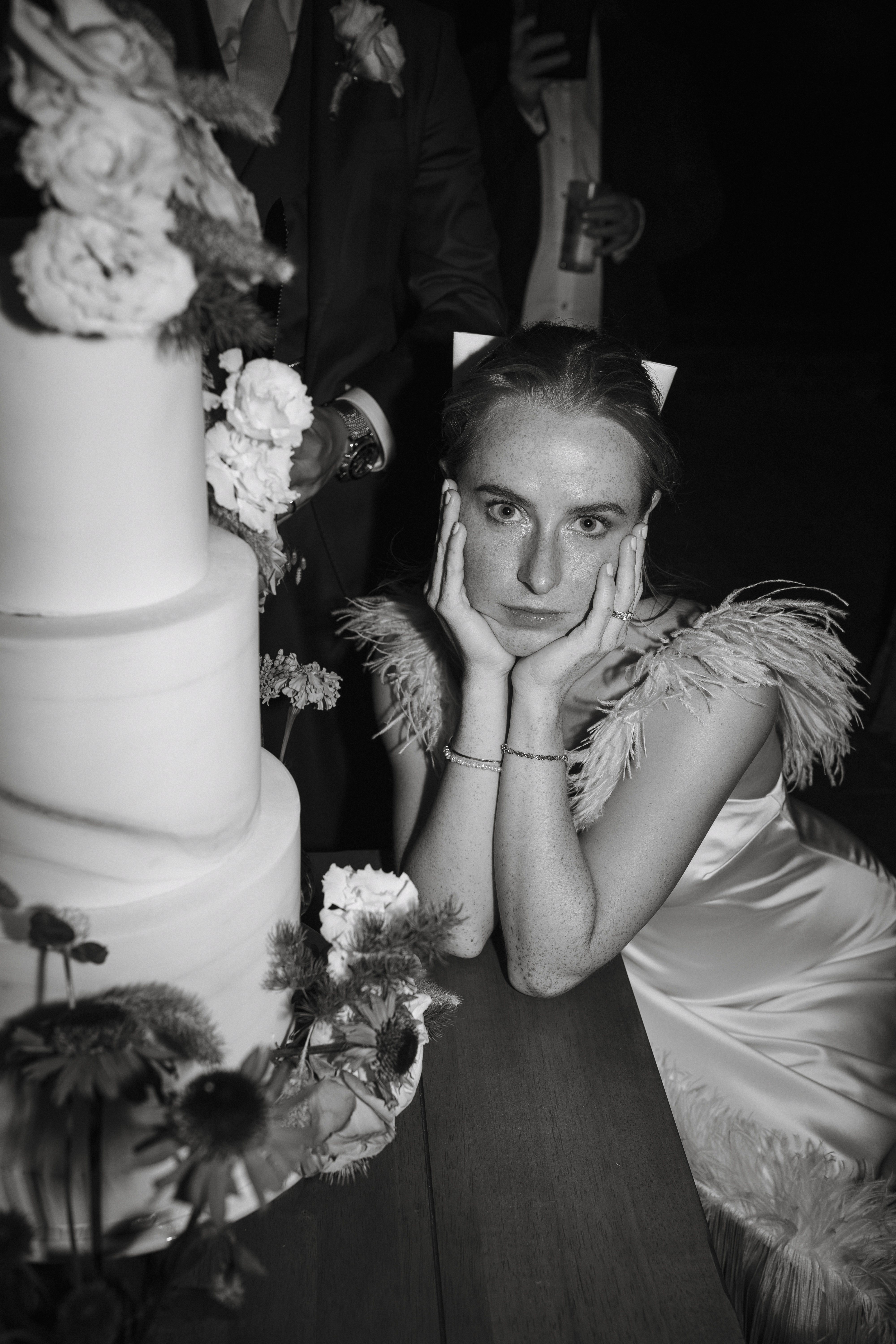 A Summer Wedding at Elmley Nature Reserve with Halfpenny London 107 Bride resting with her head. her hands next to her wedding cake. She wears an ostrich feather dress by Halfpenny London.