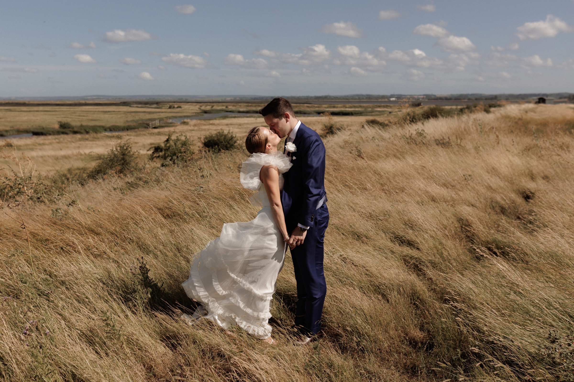 A Summer Wedding at Elmley Nature Reserve with Halfpenny London 57 Elmley Nature Reserve wedding. Image of the bride and groom kissing after the ceremony with the beautiful views and landscape in the background.