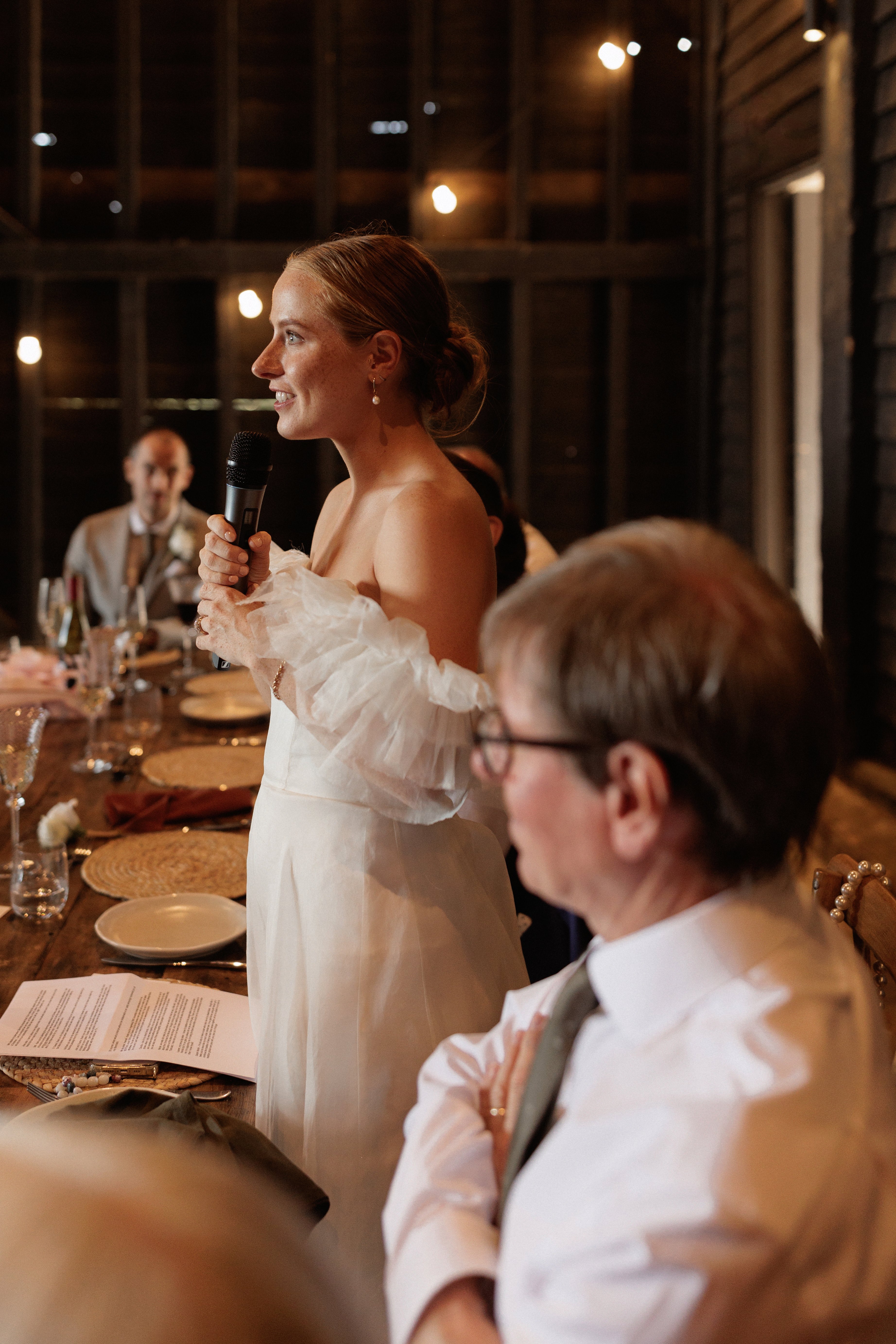 A Summer Wedding at Elmley Nature Reserve with Halfpenny London 97 Bride holding a microphone. giving a speech during a wedding reception. She wears Halfpenny London. Elmley Nature Reserve wedding venue, Kent.