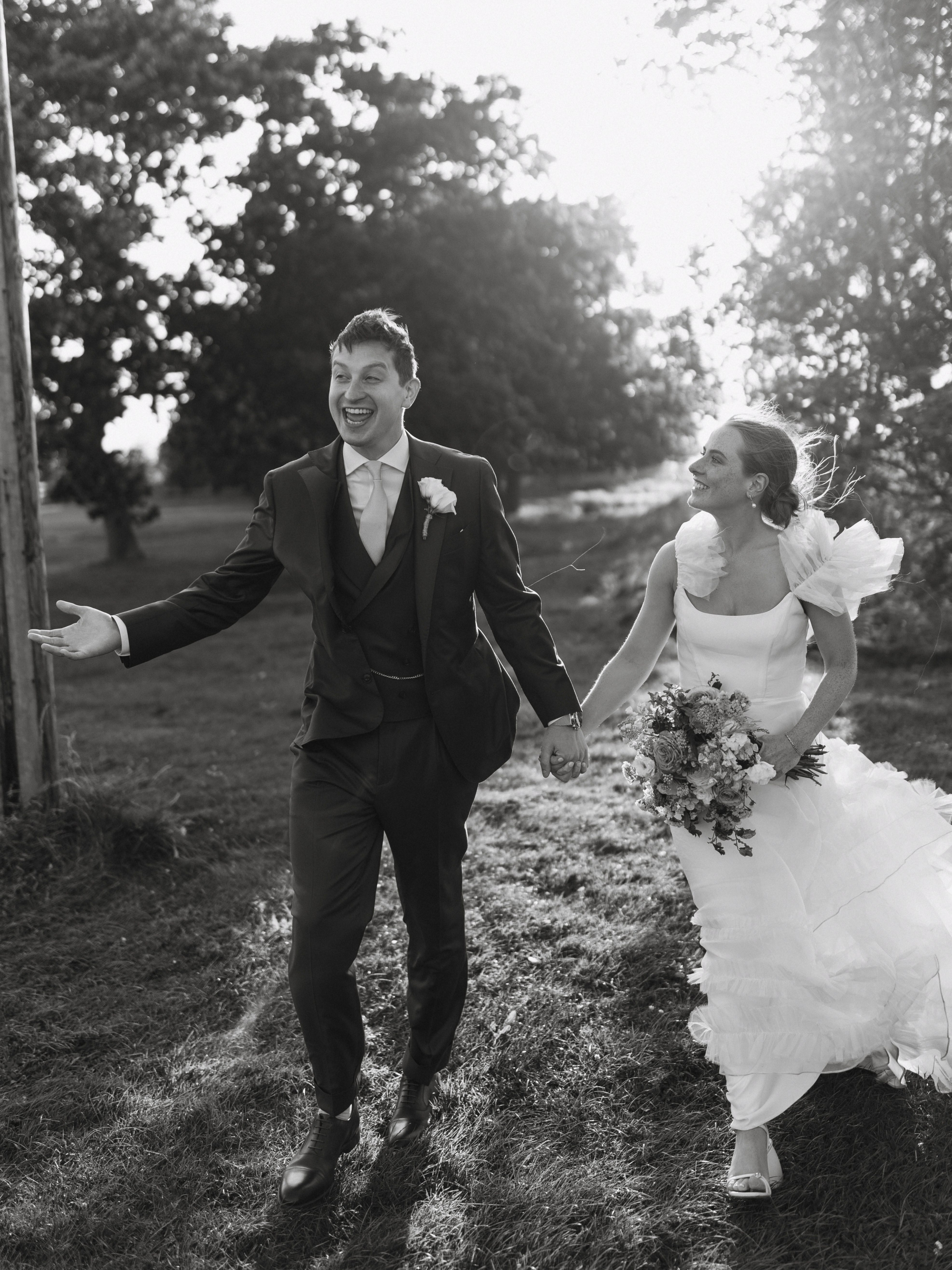A Summer Wedding at Elmley Nature Reserve with Halfpenny London 100 Black and white golden hour wedding photography at Elmley Nature Reserve in Kent. Bride wears Halfpenny London wedding dress with frilly sleeves and groom wears a dark blue 3 piece suit. They. walking, smiling. holding hands.and
