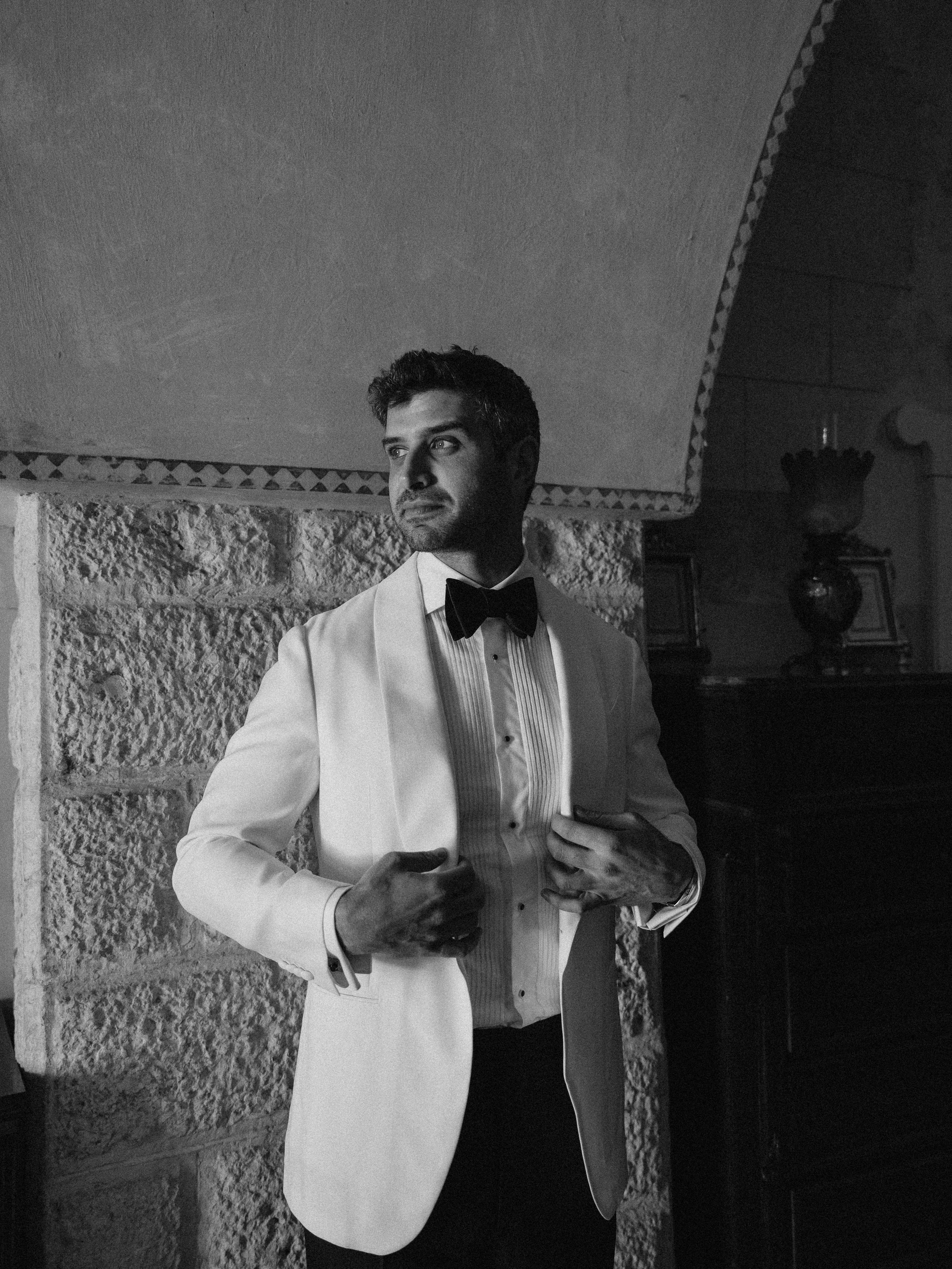 Will & Heather's Romantic Wedding in Puglia Inspired by Classic Italian Design 37 Groom getting ready at Masseria Salamina wedding venue in Puglia Italy. He wears a white tuxedo jacket and black bow tie.