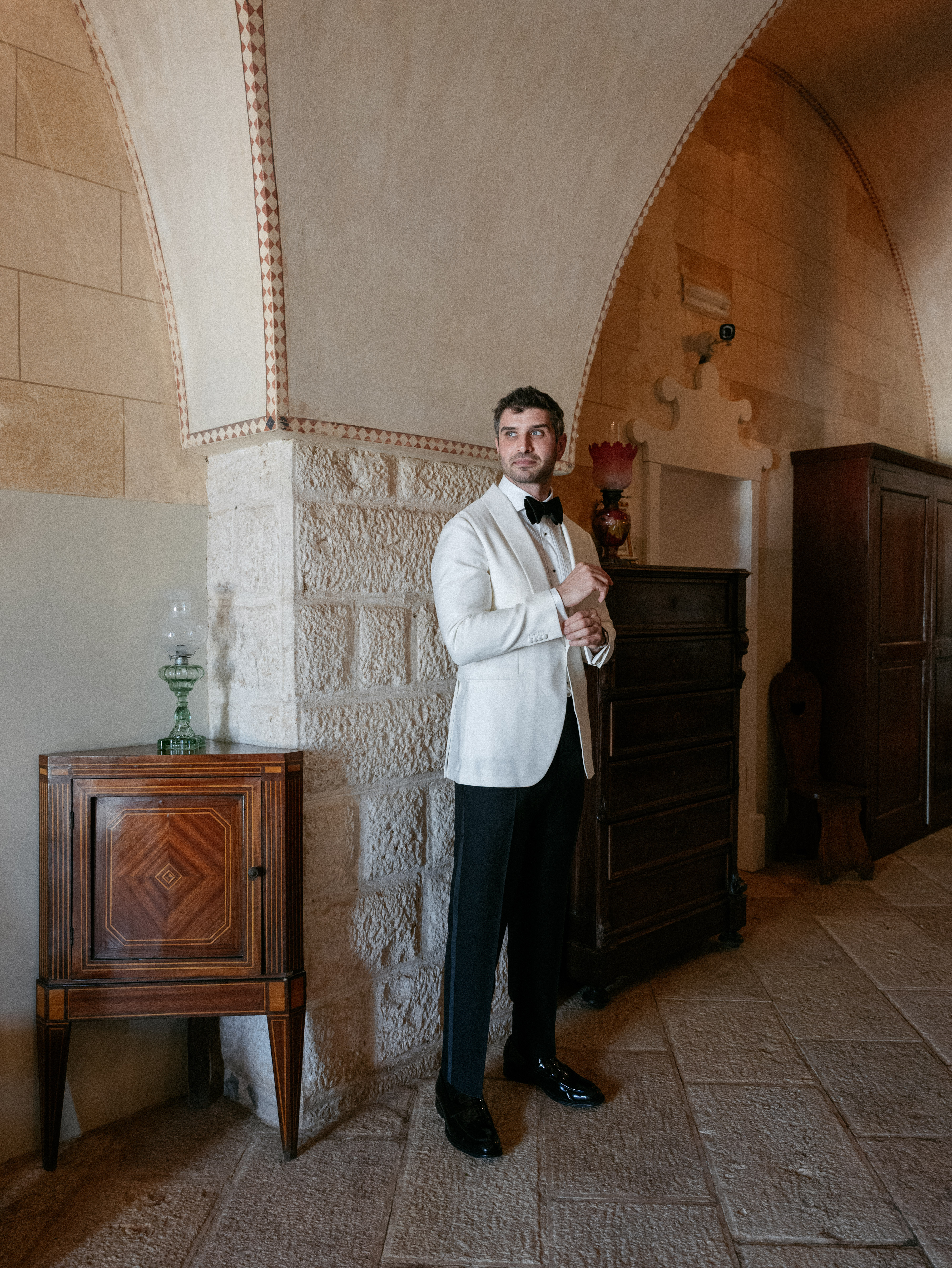 Will & Heather's Romantic Wedding in Puglia Inspired by Classic Italian Design 36 Groom getting ready at Masseria Salamina wedding venue in Puglia Italy. He wears a white tuxedo jacket and black bow tie.