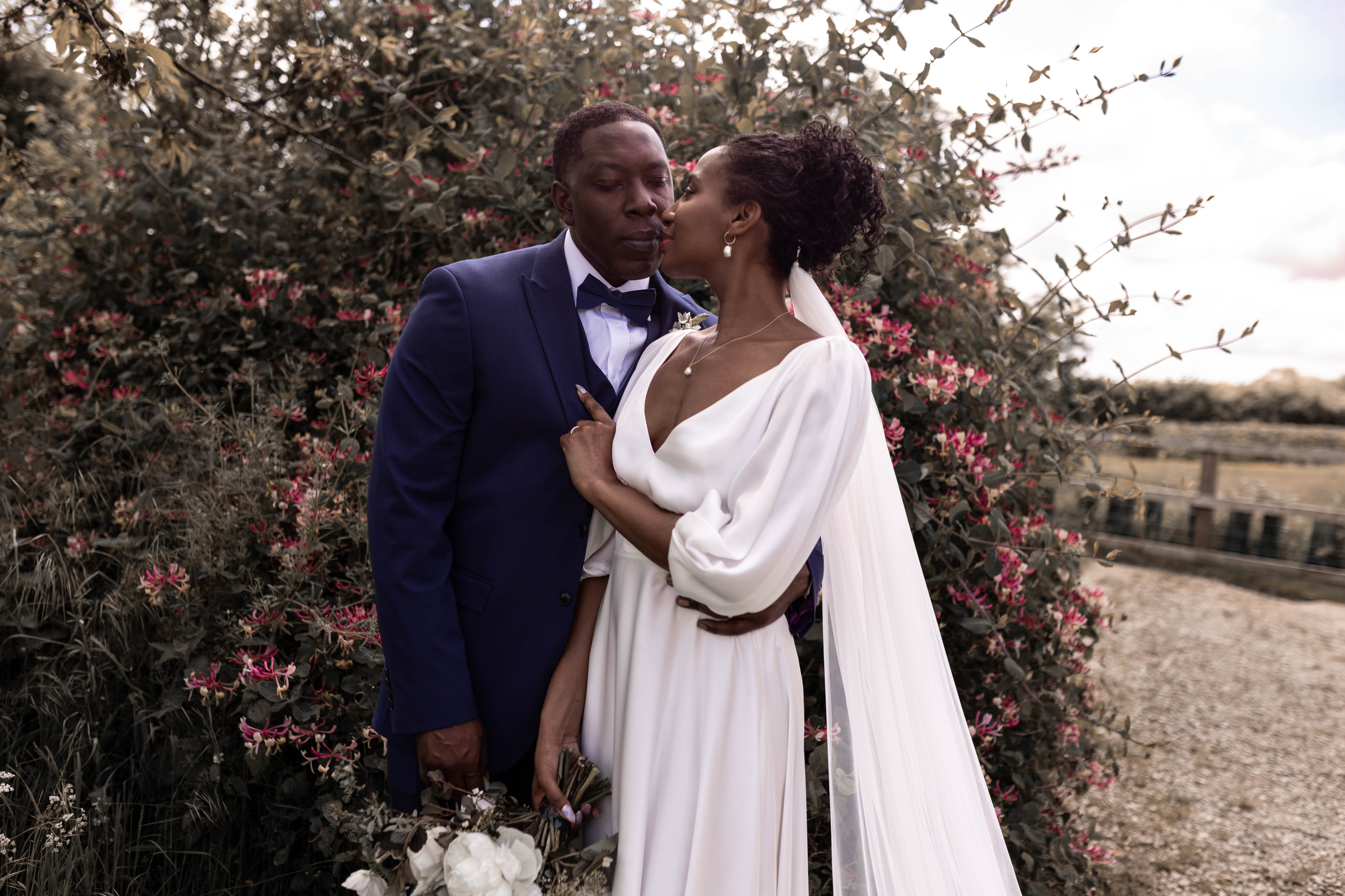 Beautiful Black bride wearing Rolling in Roses and tenderly kissing her groom's face. He wears a dark suit with a bow tie.