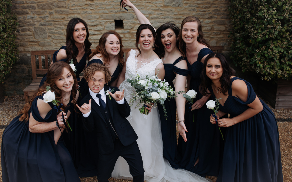 Bride surrounded by her bridesmaids in navy blue dresses and single stem white roses. Tythe, an Oxfordshire barn wedding venue. 