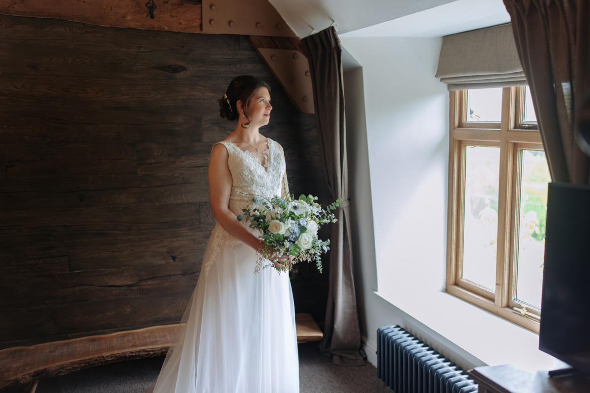 Bride holding her wedding bouquet (white and blue flowers) and looking out through the window. Wedding at Tythe, an Oxfordshire barn wedding venue. 