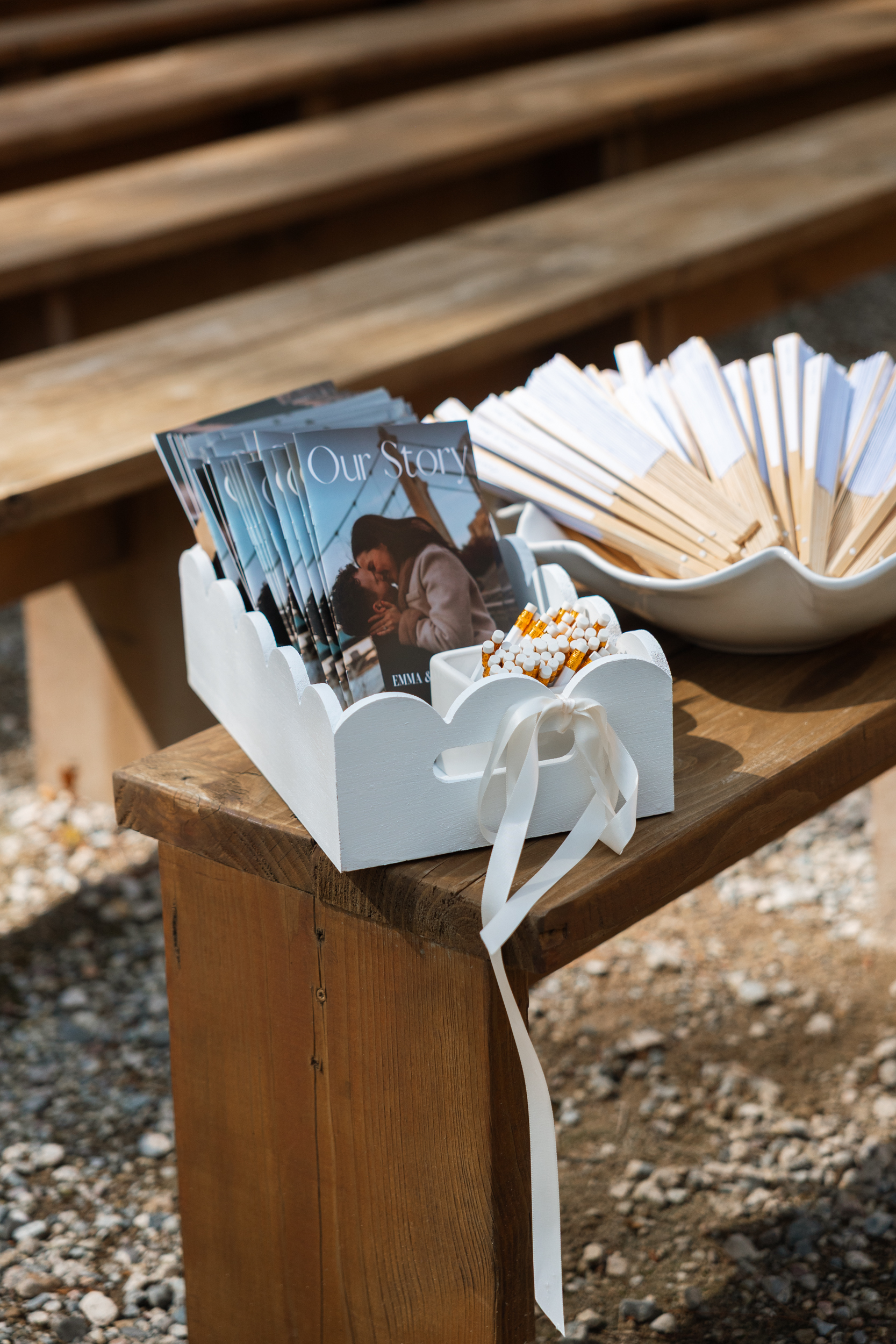 White wooden scalloped tray holding 'Our Story' booklets for wedding guests. Nearby, a bowl containing fans.