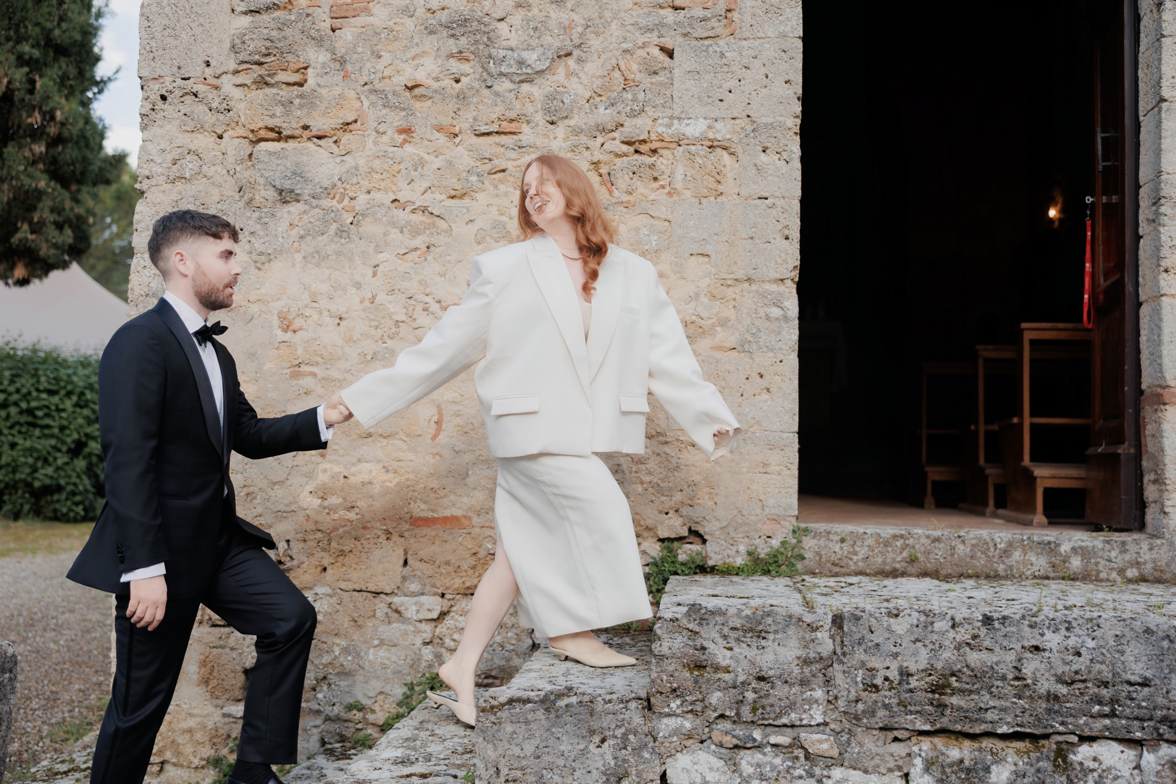 An Arakii Suit with Oversized Jacket for a Chic Italian Wedding at Antico Borgo 71 Bride wearing a two-piece skirt and jacket by Swedish brand Arakii and elegant Marcella NYC mules, for her destination wedding at Antico Borgo San Lorenzo in Poggibonsi, Italy. She is climbing steps and looking back over her shoulder towards her groom behind.